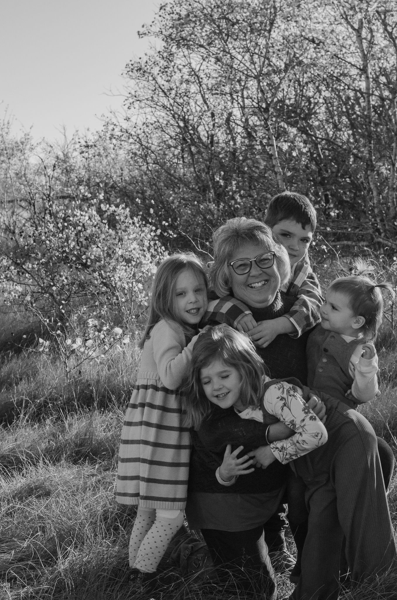 Black and white photo of a grandmother smiling and hugging four young grandchildren outdoors among blooming trees.