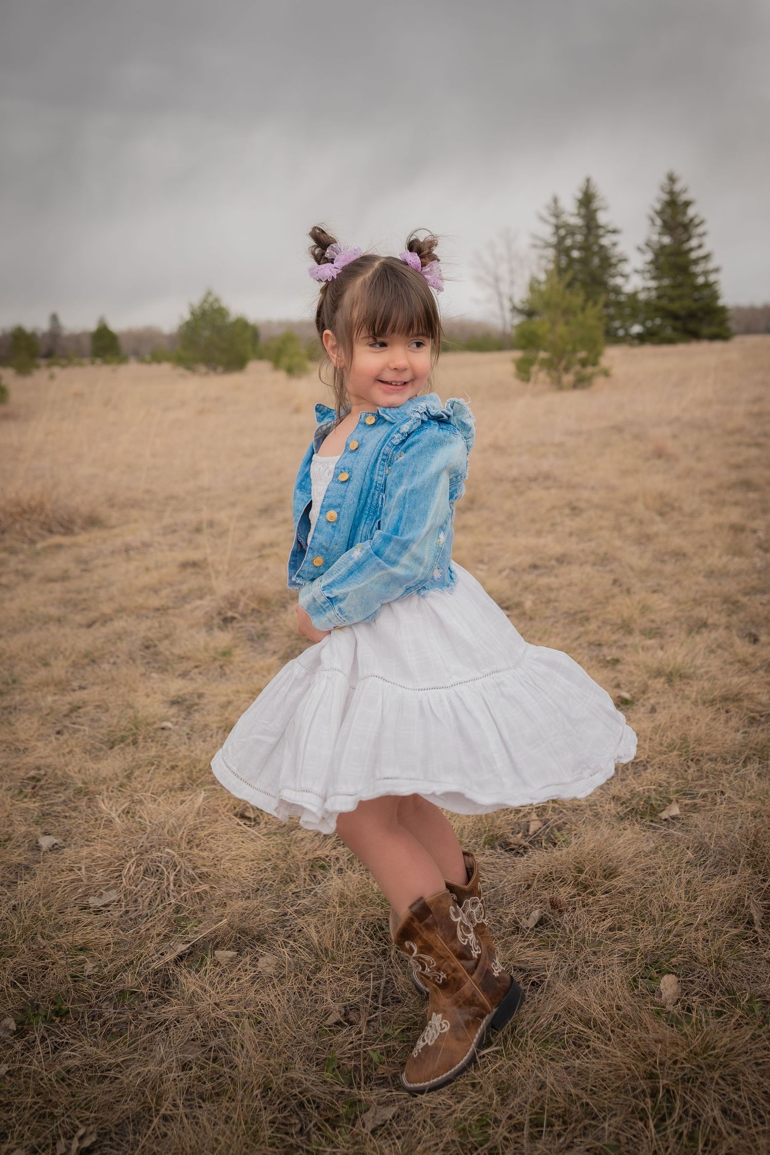 Smiling little girl in denim jacket, white skirt, and cowboy boots twirling in a dry field.