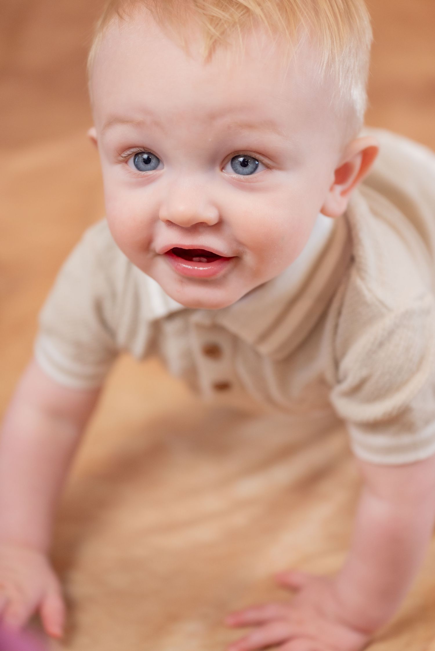 Smiling baby boy with blue eyes and blonde hair crawling on a wooden floor wearing a beige polo onesie.