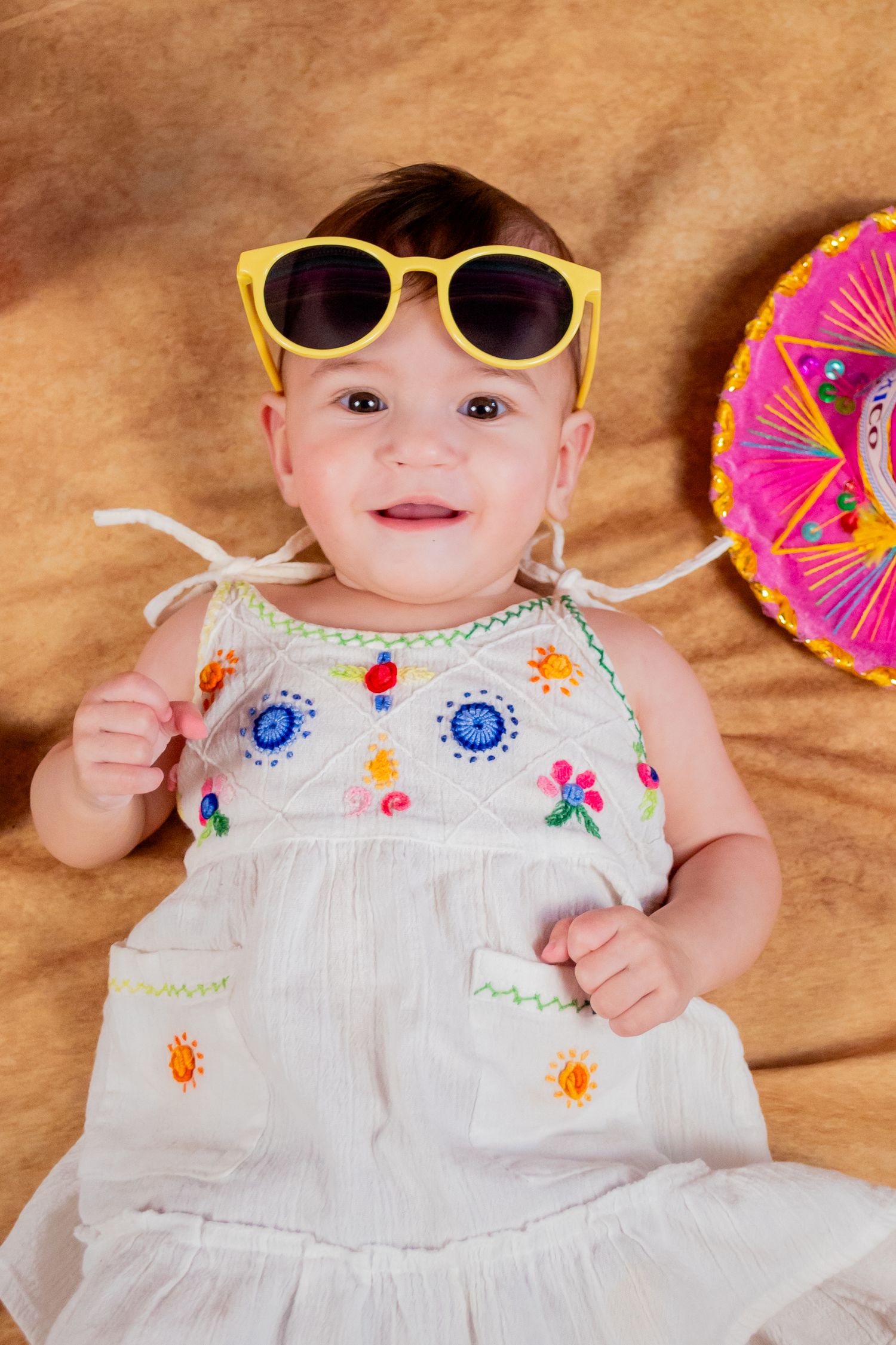 Adorable baby girl wearing oversized yellow sunglasses and embroidered white dress, smiling next to colorful sombrero.