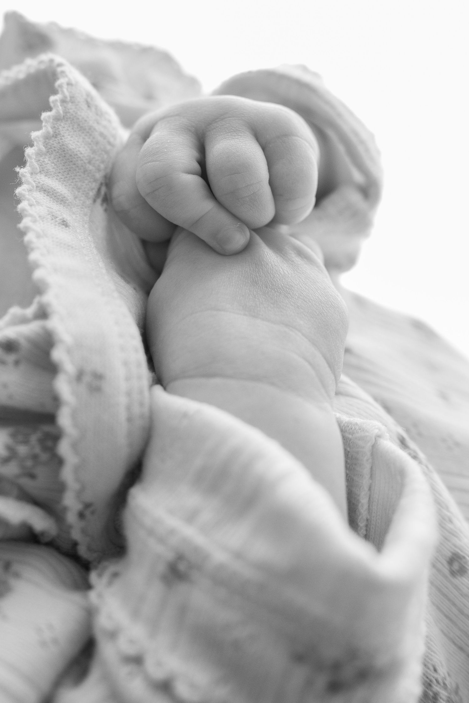 Black and white close-up of a newborn baby's tiny hand grasping softly while wrapped in a white muslin blanket.