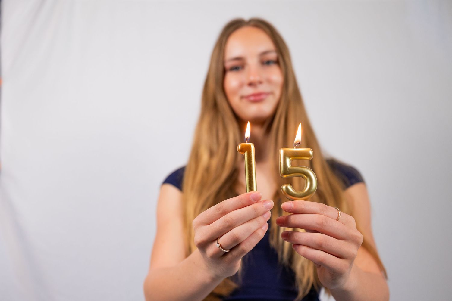 Young woman holding two lit gold number 15 birthday candles, smiling softly against a white background.