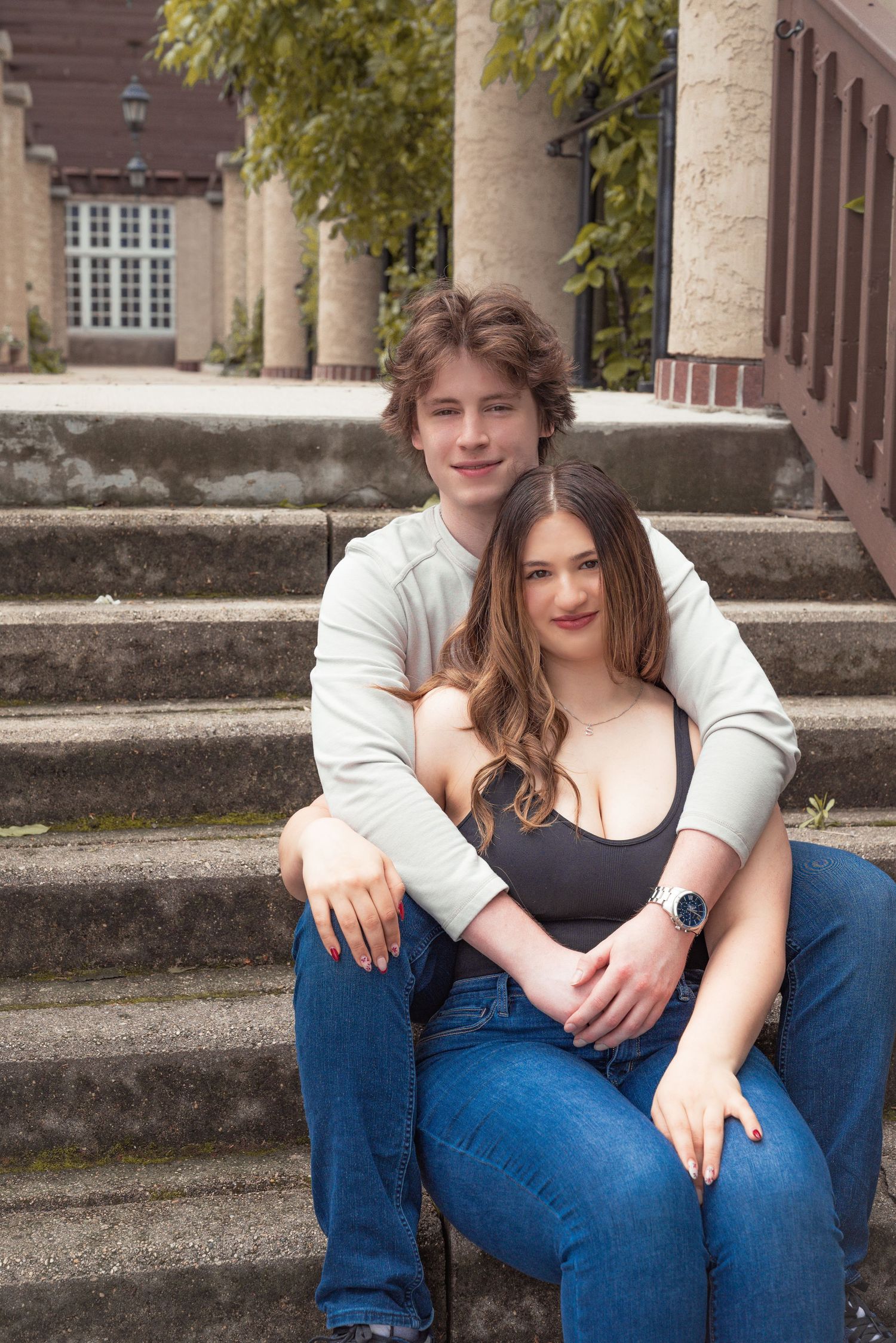 Young couple sitting on stone steps, smiling, wearing casual jeans and tops, with a classic building in the background.