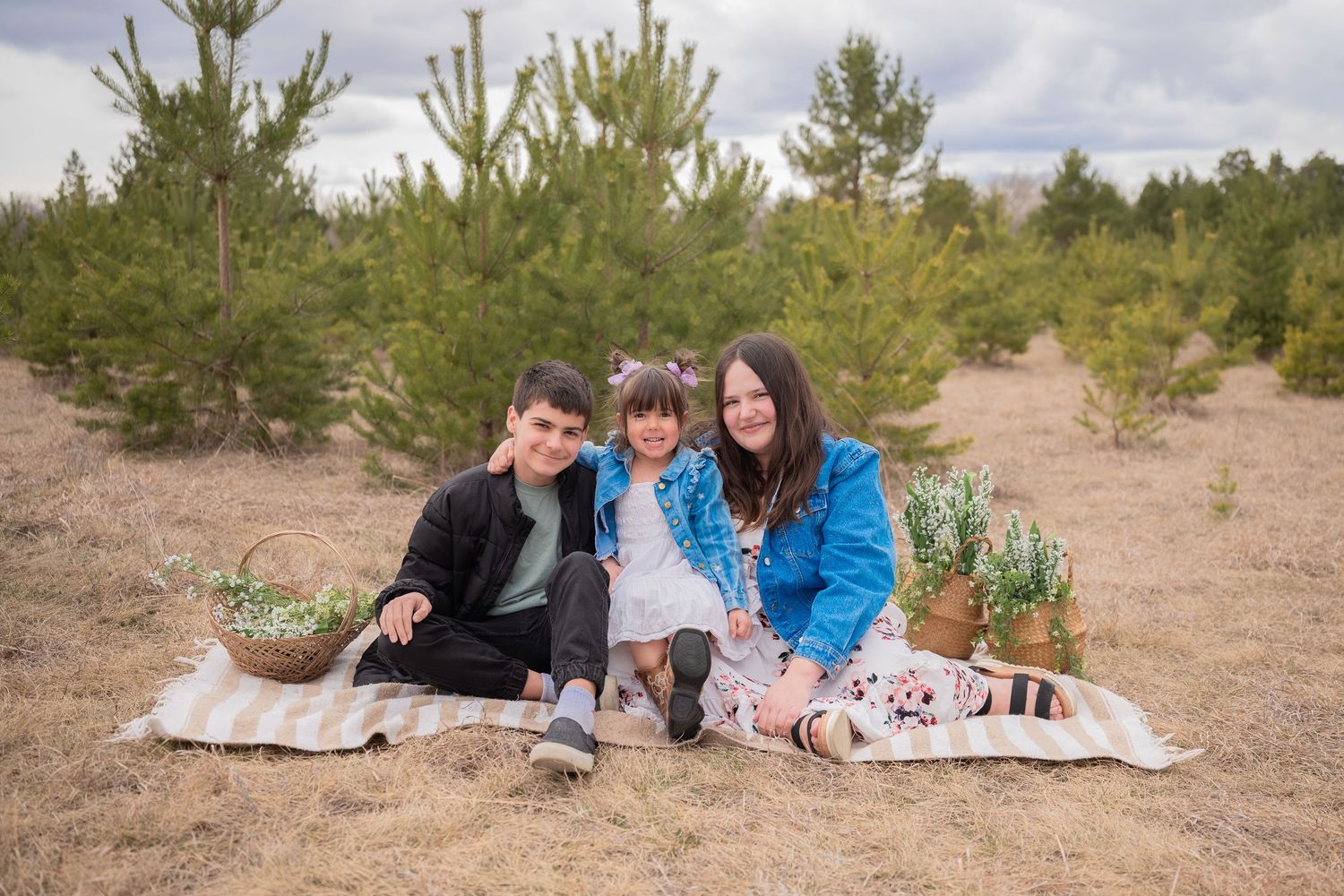 Family of three sitting on blanket in pine forest with flower baskets during outdoor picnic photo session.