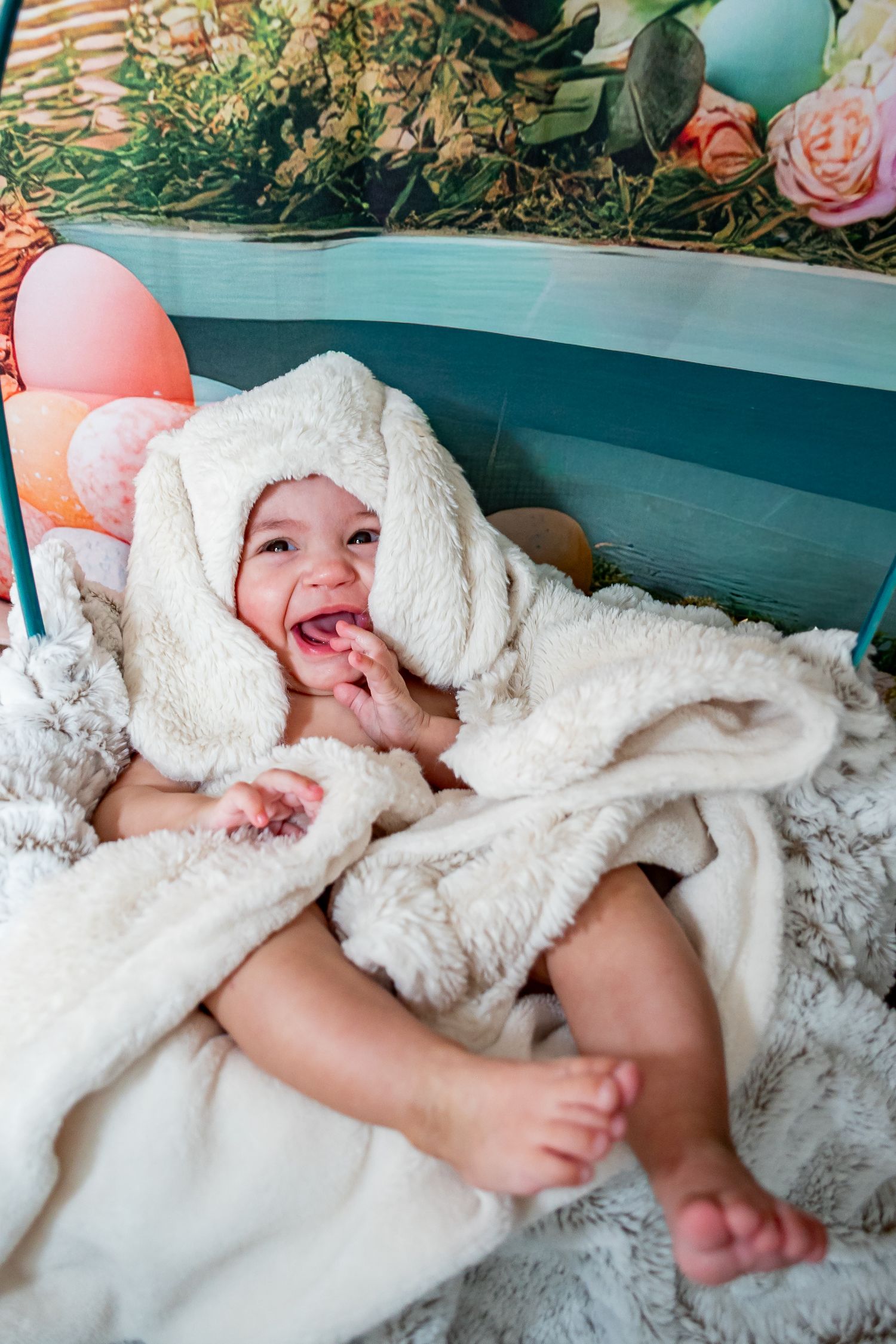 Happy baby wrapped in fluffy white bunny blanket, laughing with mouth open, surrounded by Easter-themed decorations.