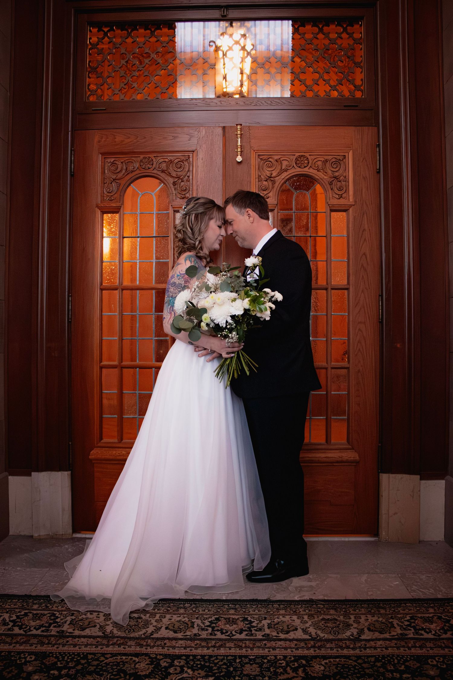 Newlyweds share an intimate moment before ornate wooden doors, bride holding a white floral bouquet.