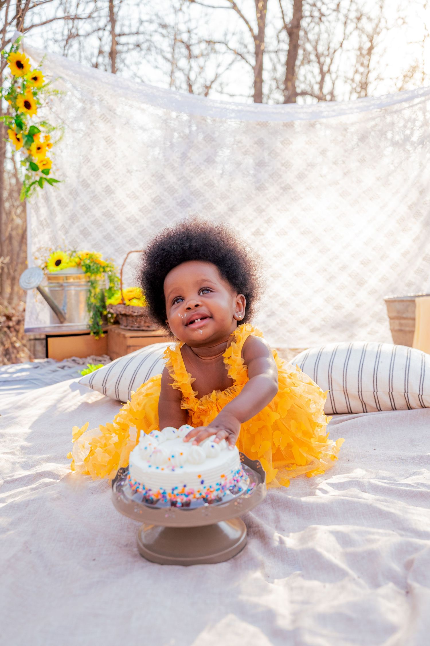 An adorable baby girl in a yellow ruffle outfit smiles beside a birthday smash cake at an outdoor party.