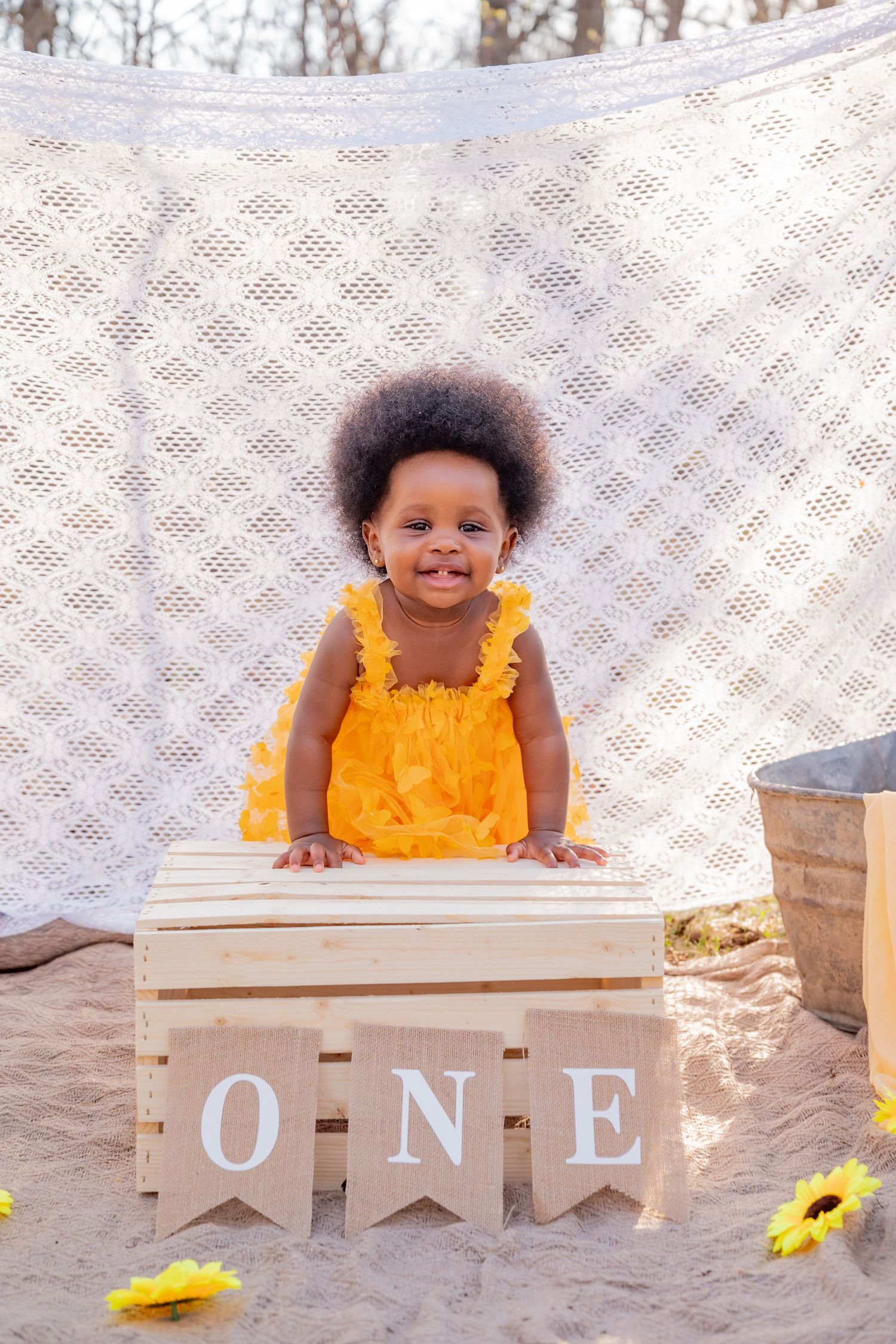 Smiling toddler girl in yellow dress standing behind wooden crate with ONE sign, first birthday photo shoot.