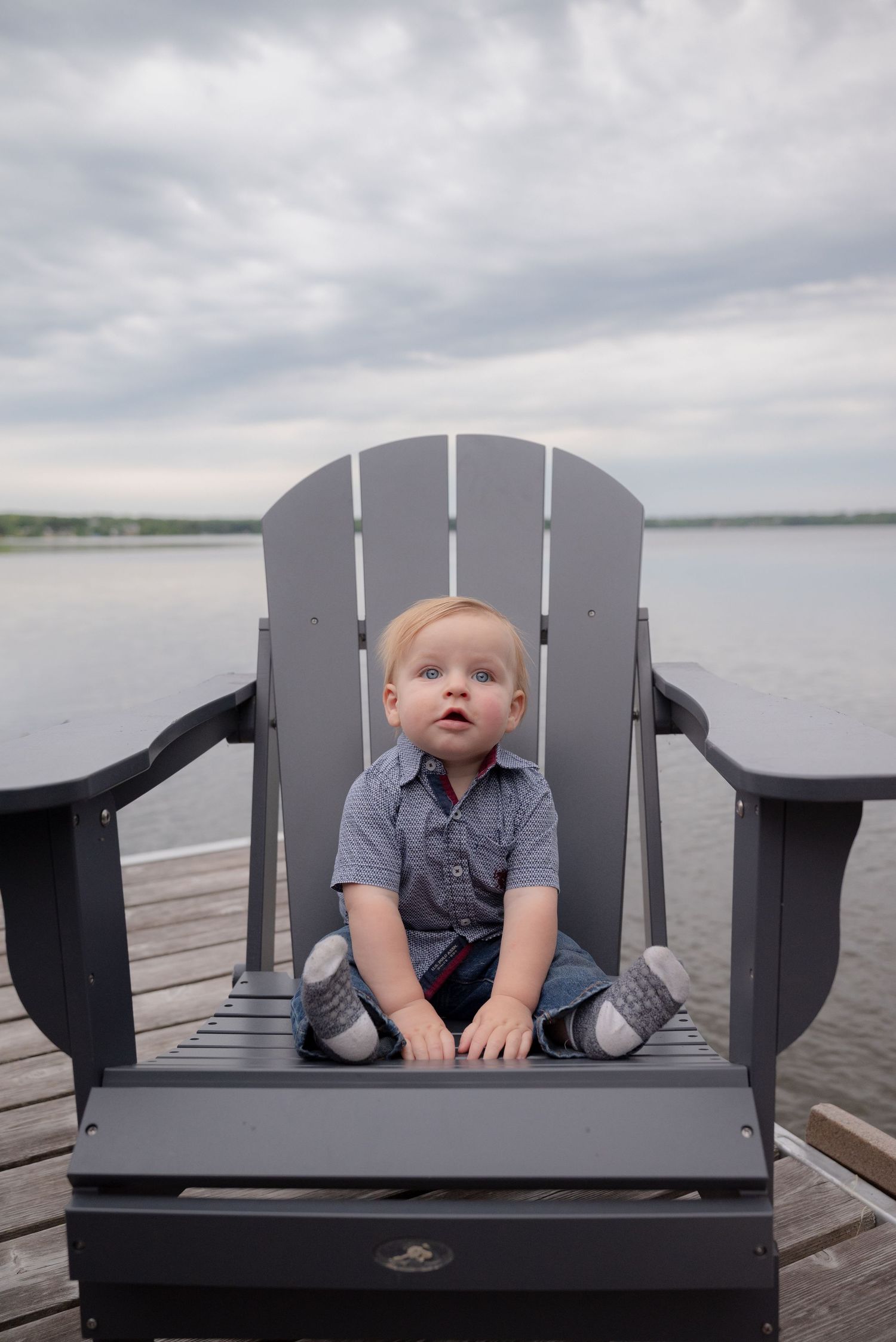 Adorable toddler sitting in a gray Adirondack chair on a dock by a lake under a cloudy sky.