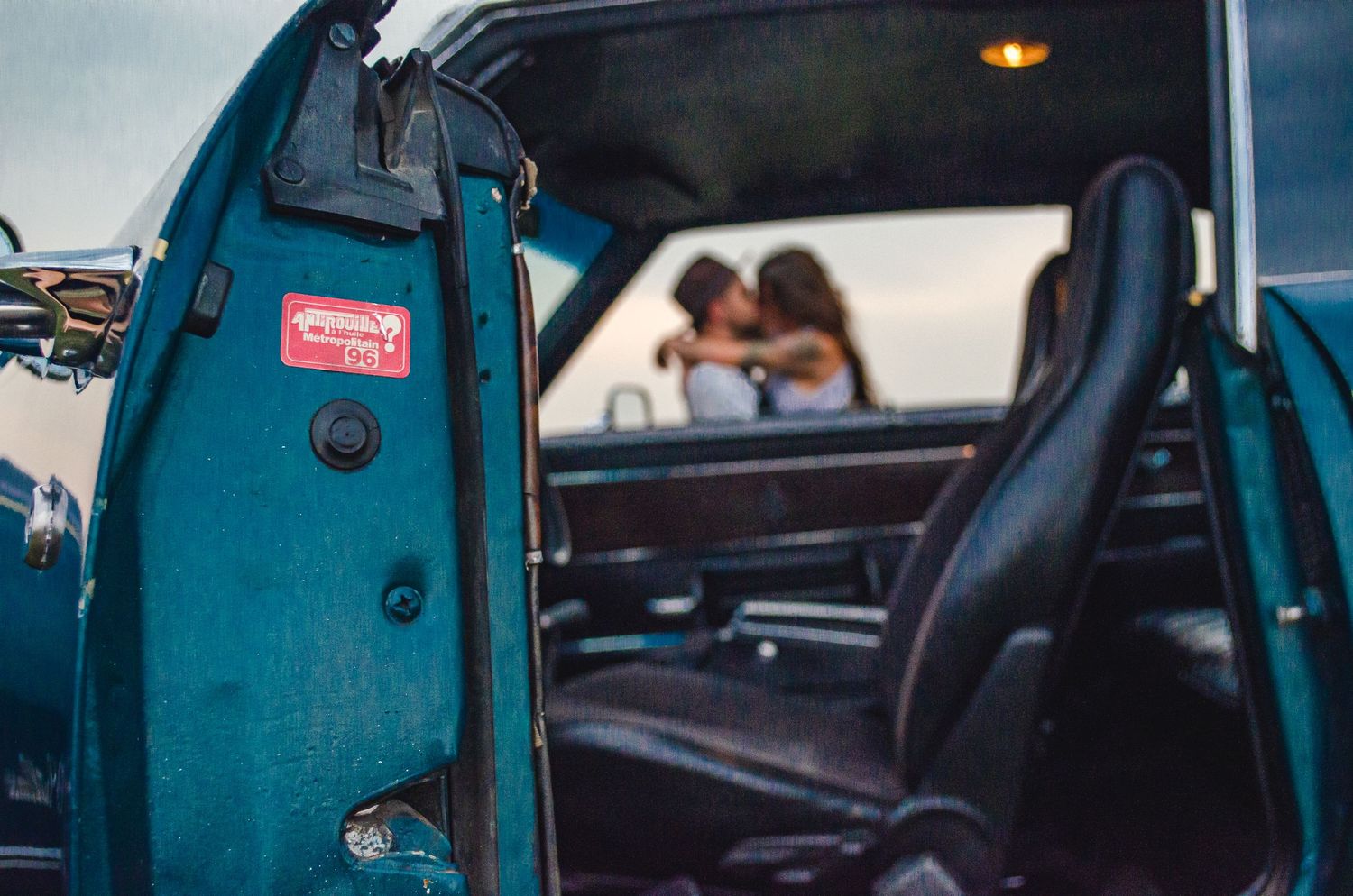 Couple sharing a romantic kiss in a vintage teal classic car with black interior seats.