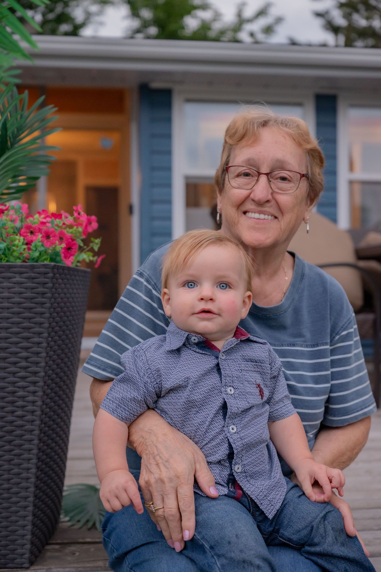 Elderly woman smiling and holding a blue-eyed baby boy on a wooden deck with pink flowers and blue house backdrop.