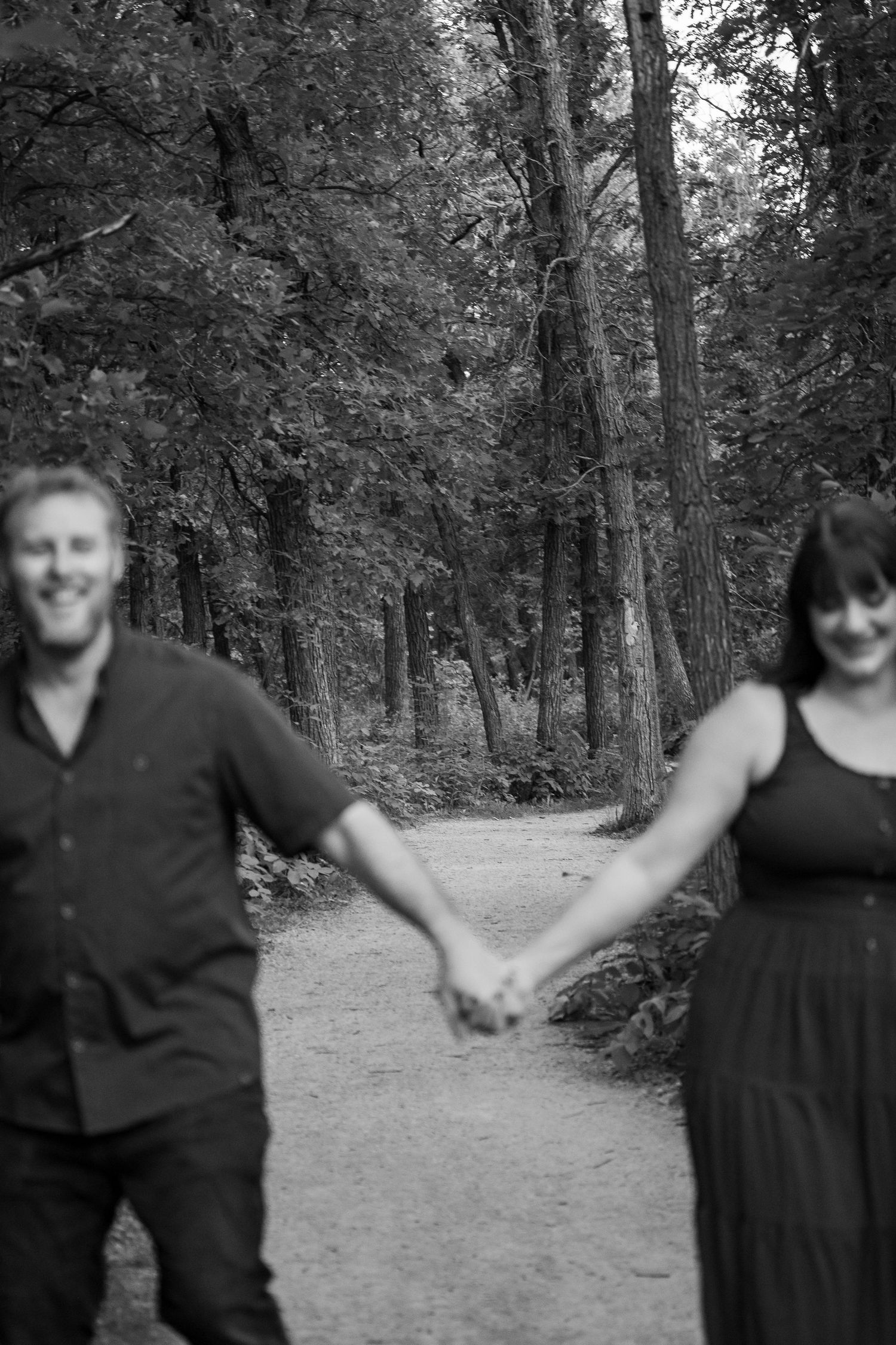 Black and white photo of a smiling couple holding hands on a forest path surrounded by tall trees.