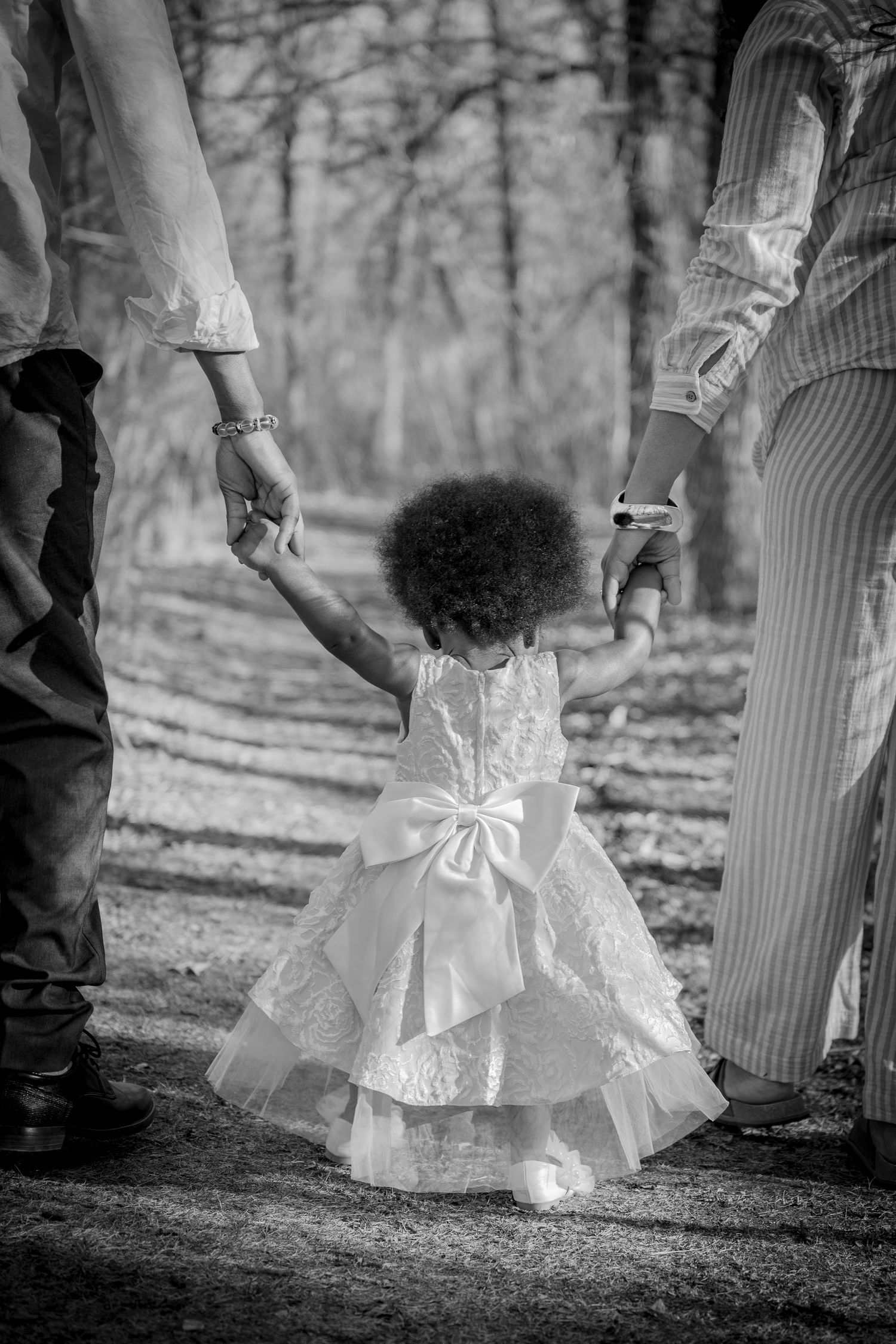 Black and white photo of toddler girl in white dress holding parents hands outdoors.