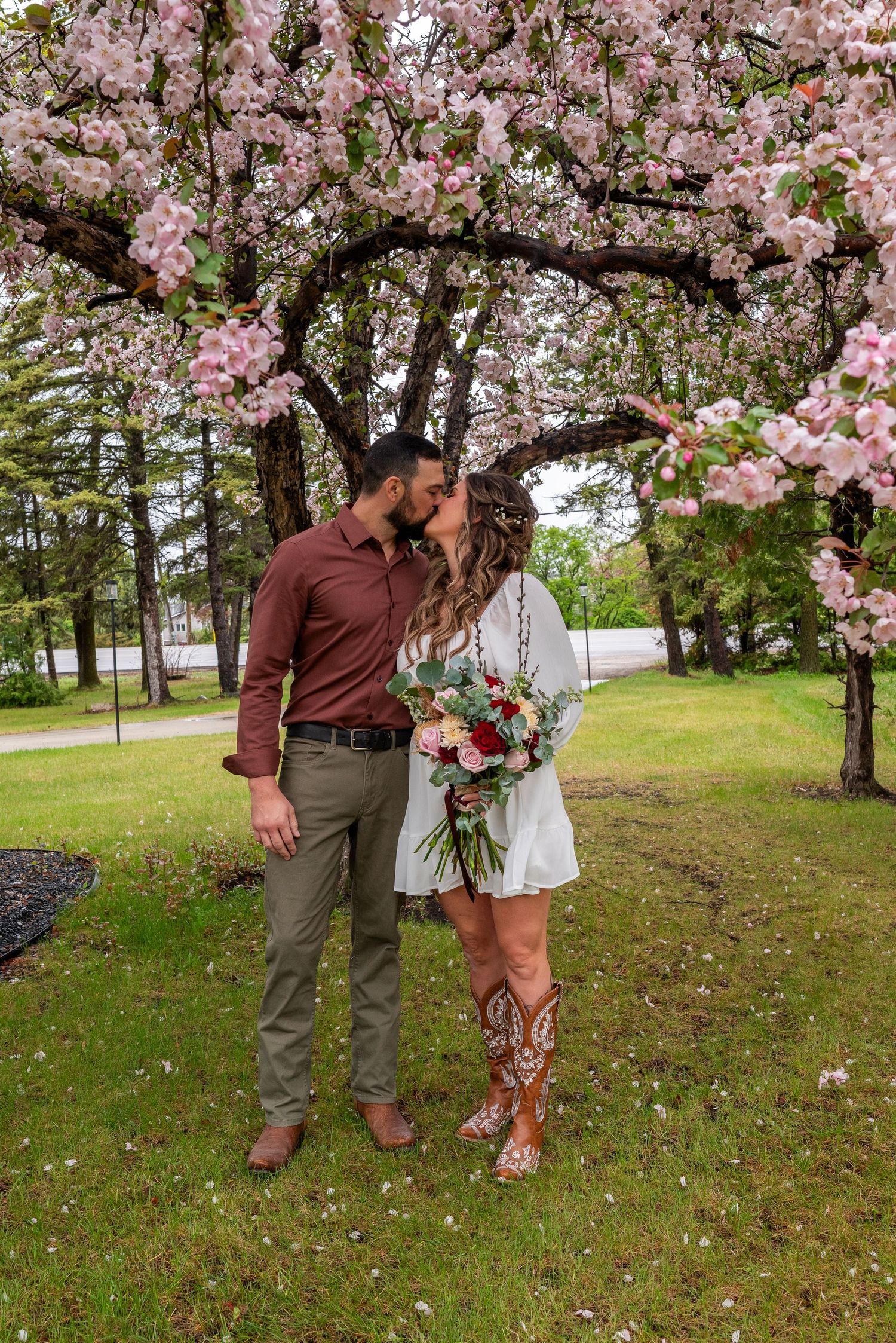 Couple kissing under pink cherry blossom tree, bride in white dress and cowboy boots holding bouquet, groom in brown shirt.