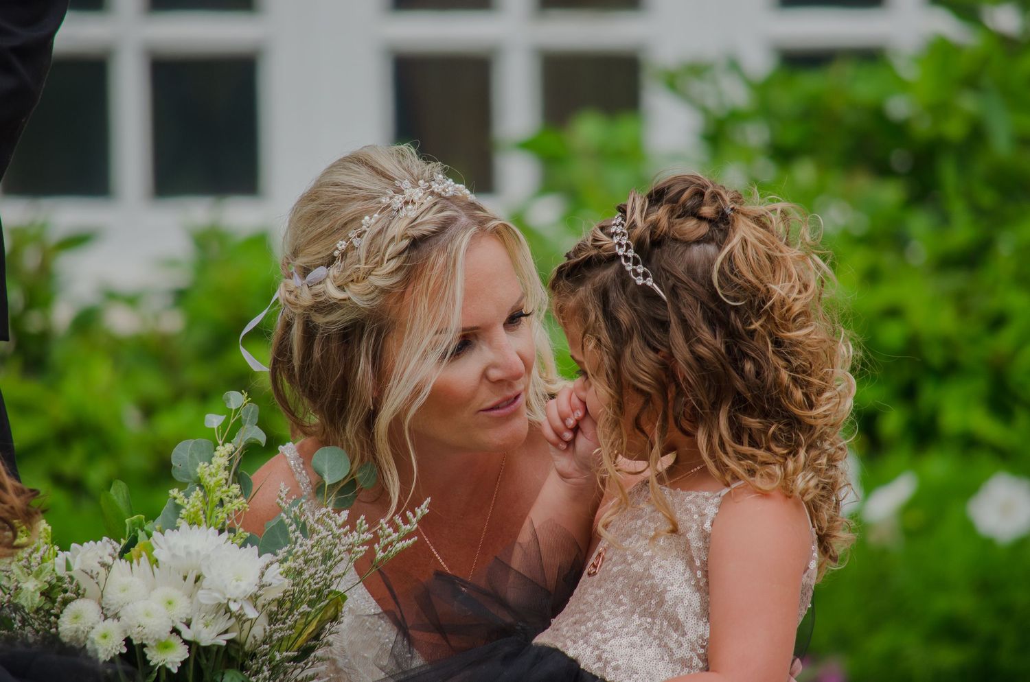 Bride in wedding dress with floral crown smiles tenderly at curly-haired flower girl outdoors.