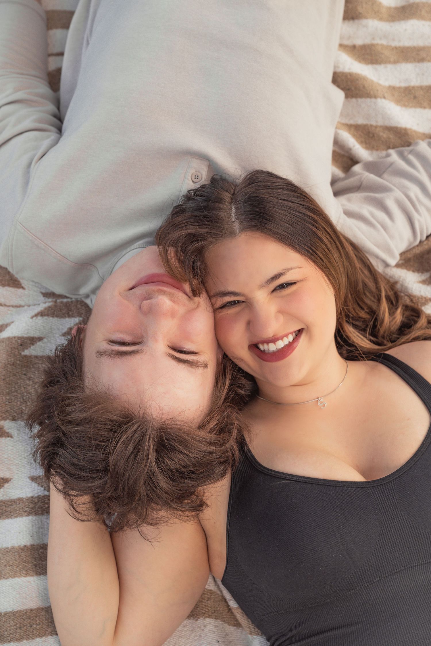 Two young people smiling and laughing while lying on a striped blanket, heads together in a joyful moment.