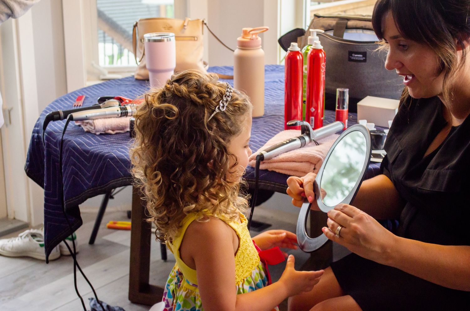 A hairstylist shows a young girl with curly hair her reflection in a mirror during a salon styling session.