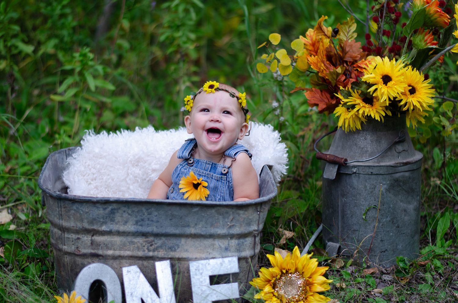 Happy baby in overalls sitting in a rustic metal tub with sunflowers and fall foliage for a first birthday photo.