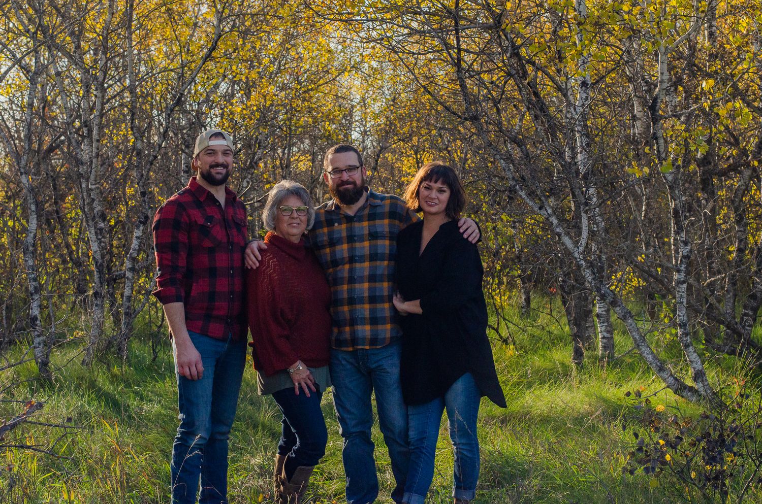 Four people smiling together in a golden autumn forest, wearing casual flannel and dark clothing for a family portrait.