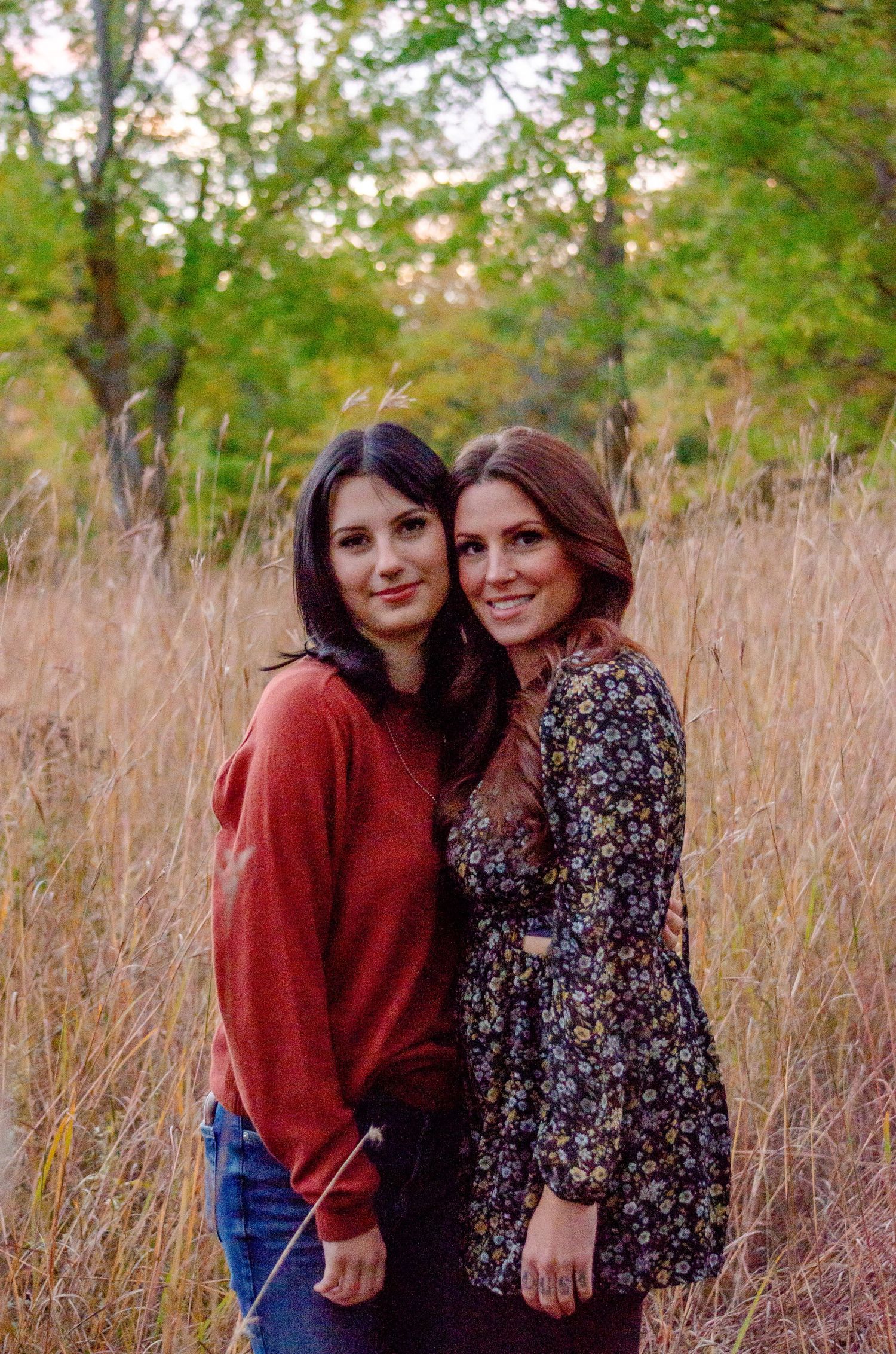Two young women smiling and posing together in a golden field with autumn trees in the background.