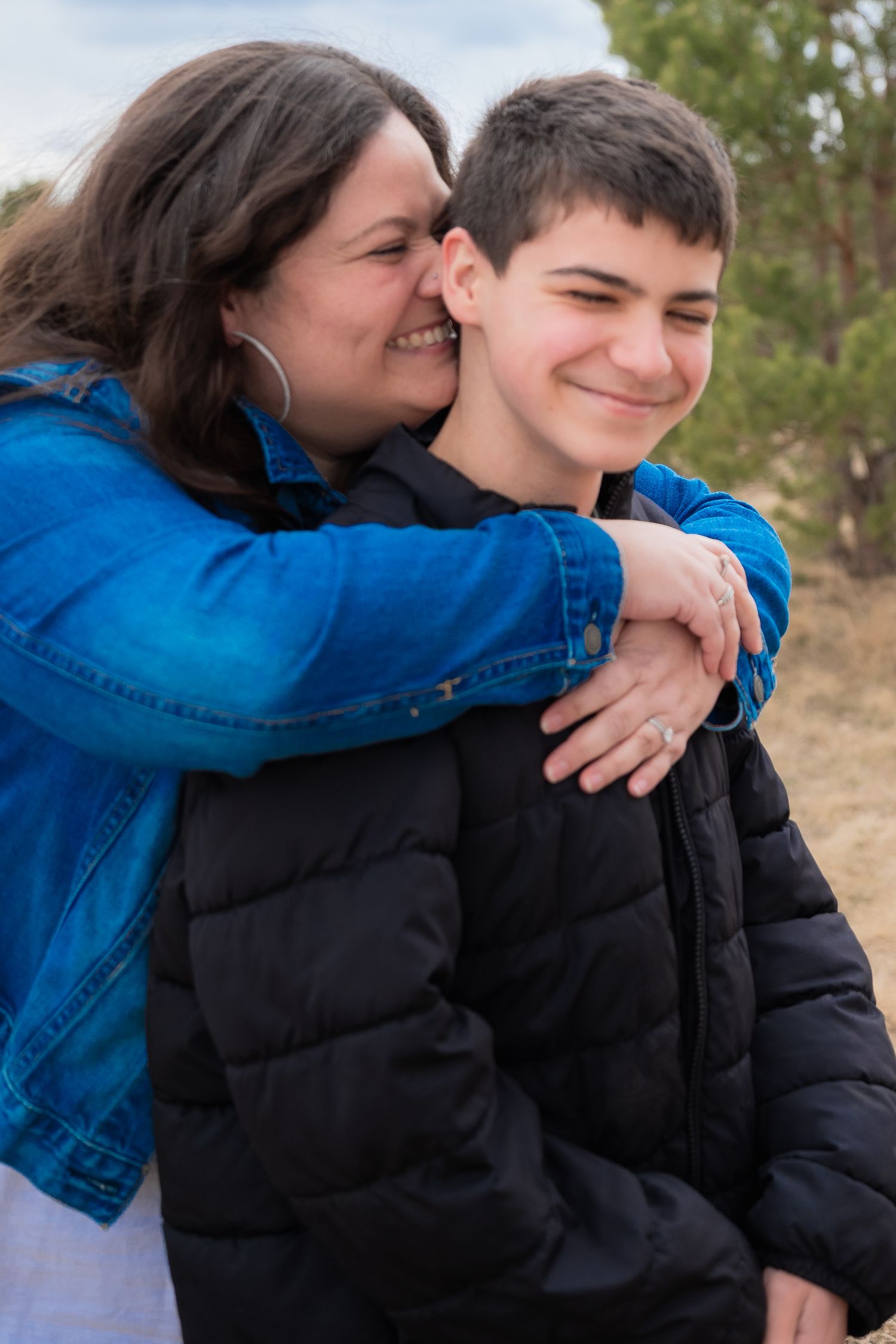 A smiling mother in a blue denim jacket hugs her teenage son outdoors in a natural setting.