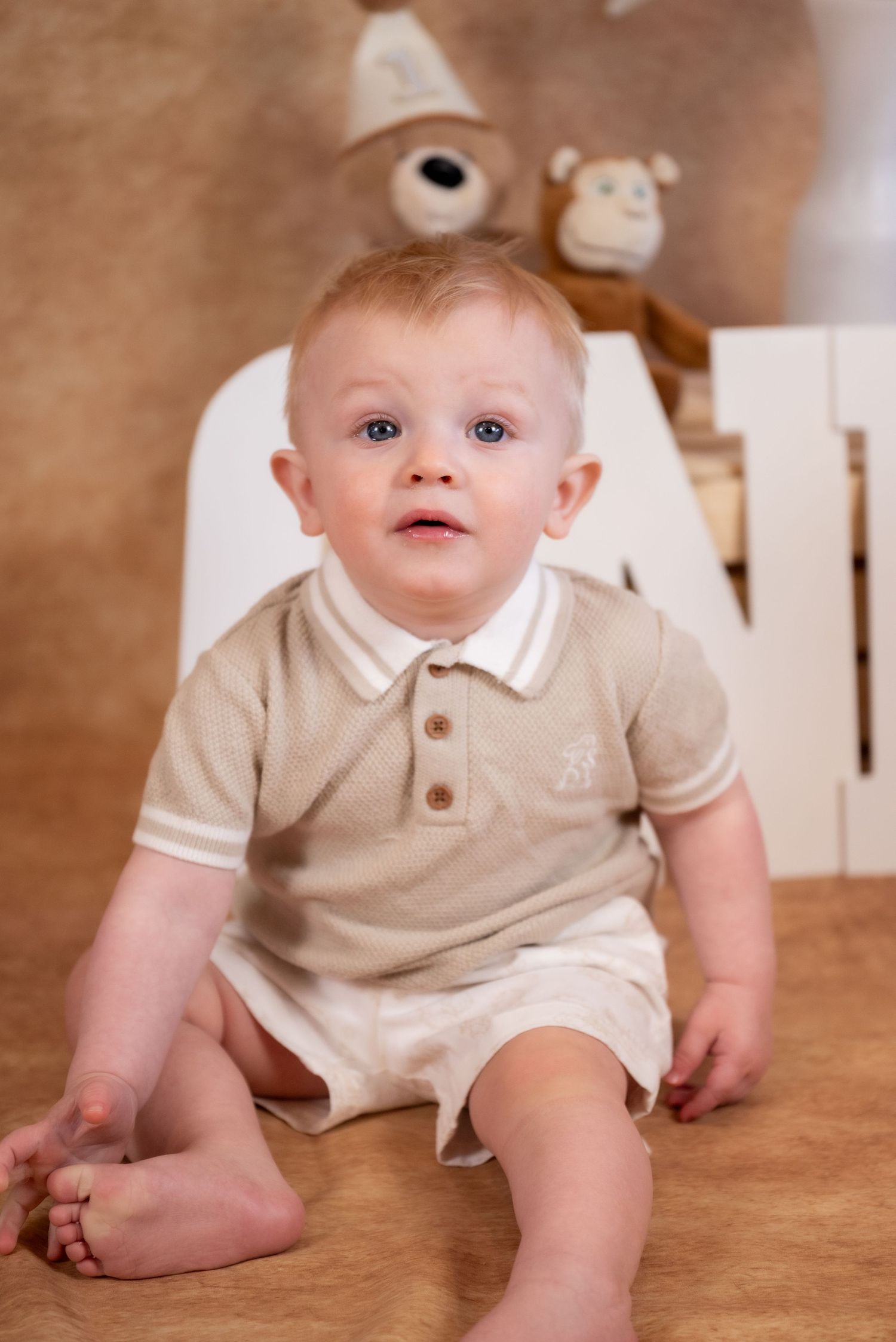 Adorable baby boy in beige knit polo romper sitting on floor with teddy bears in background.