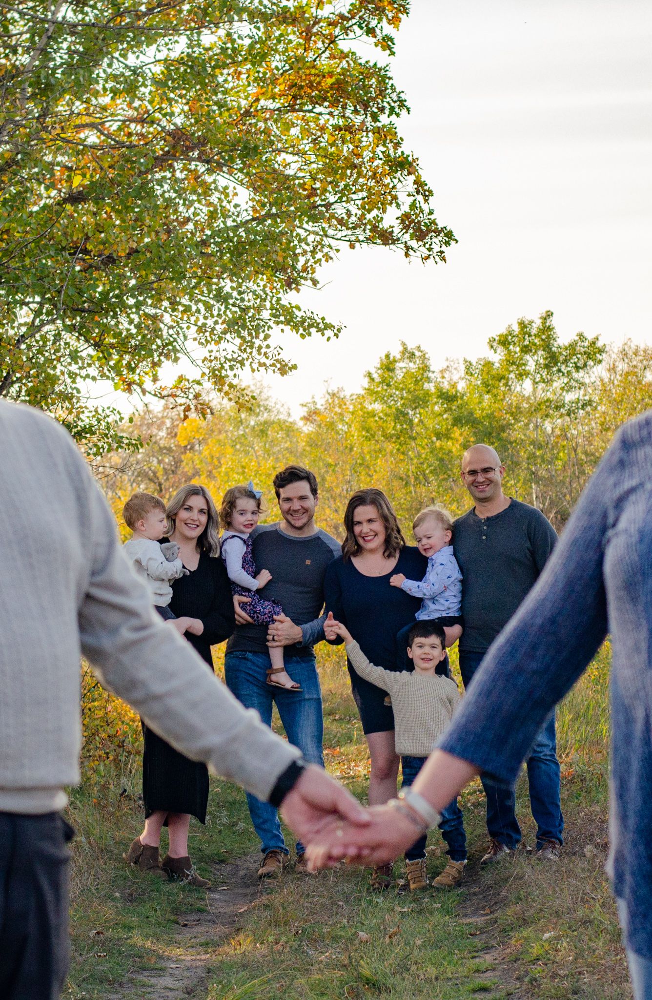 Extended family gathering outdoors in fall foliage, holding hands and smiling on a nature path.