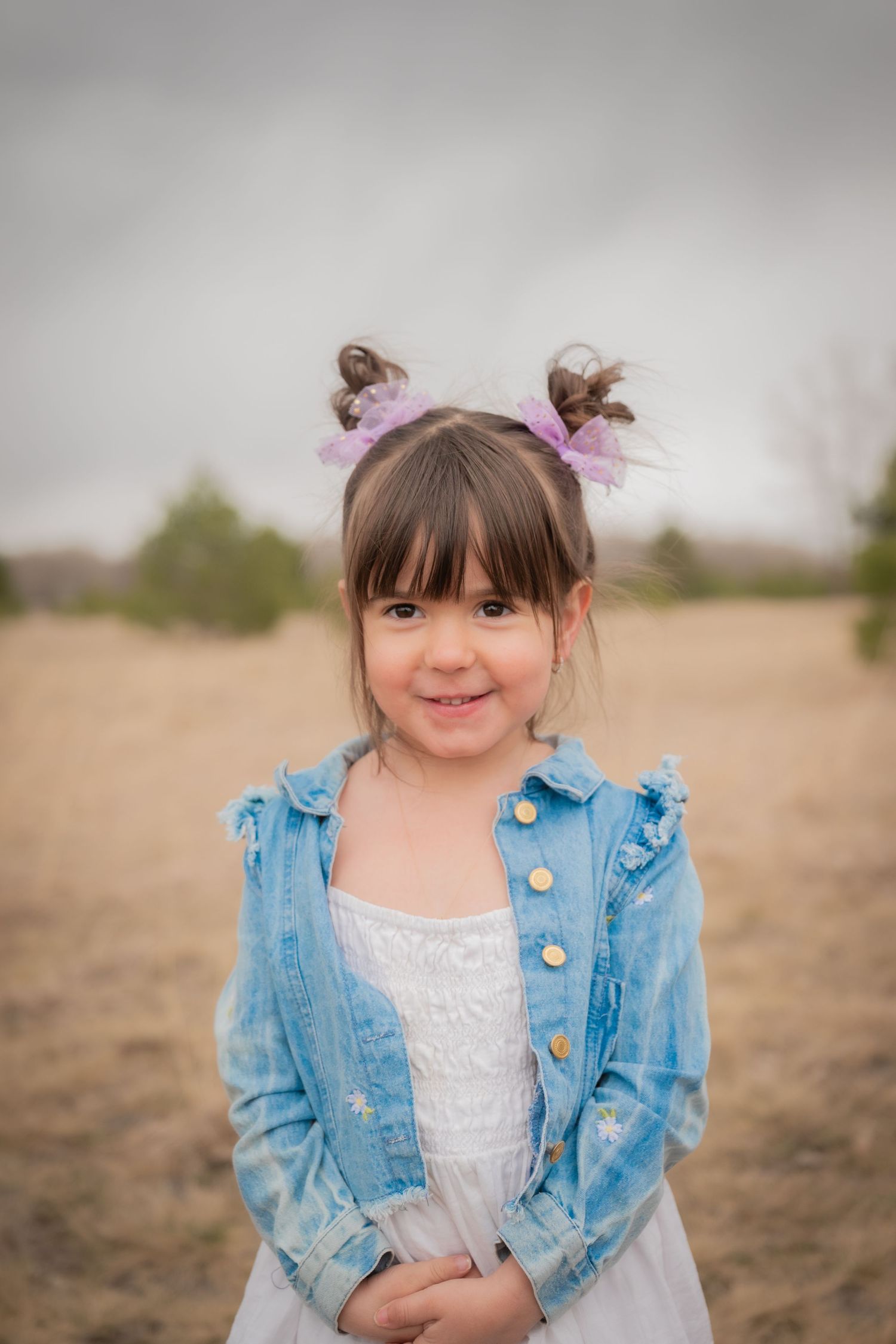 A young girl with pigtails and purple bows wearing a denim jacket over a white dress smiles outdoors.