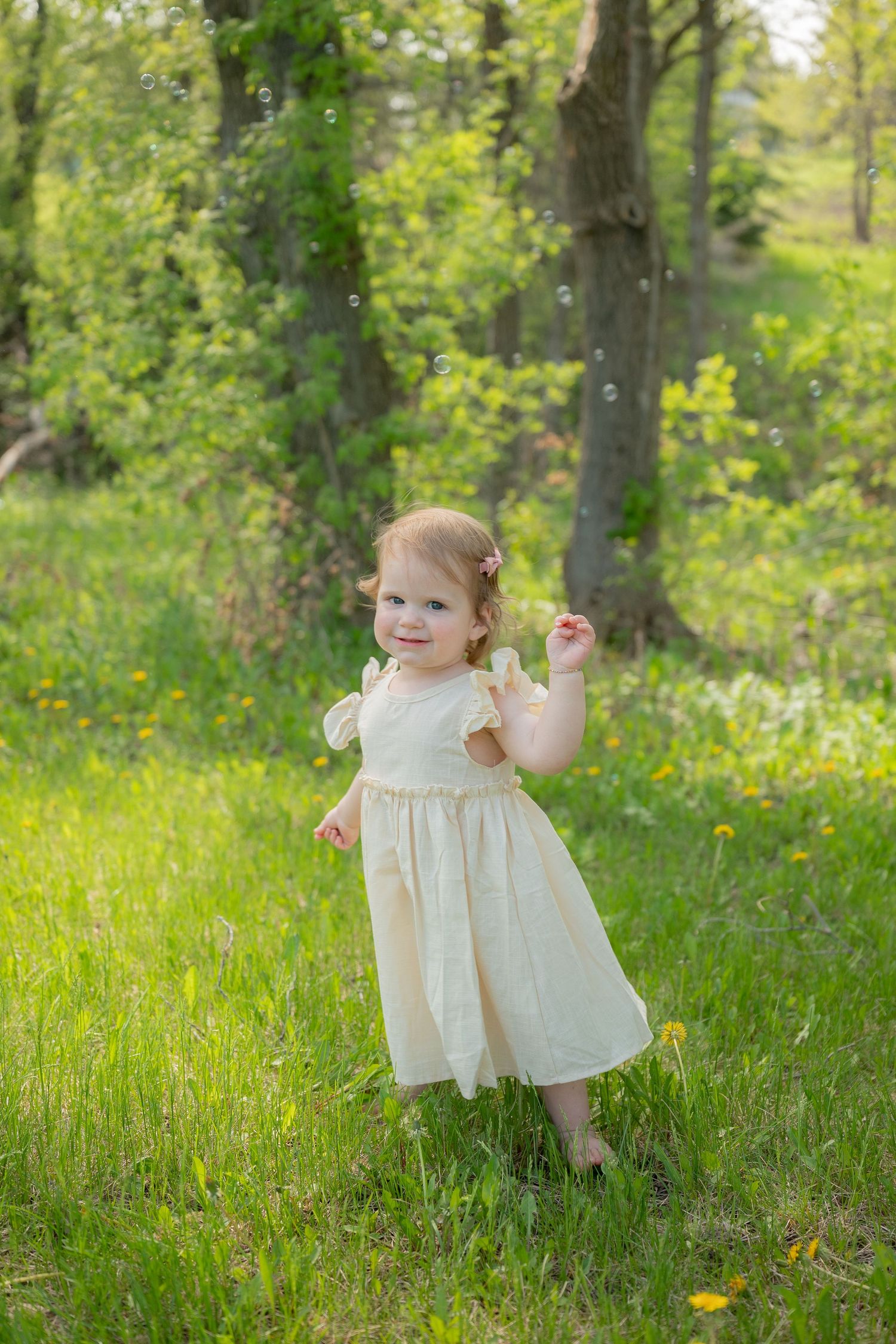 Toddler girl in cream dress playing with bubbles in a lush green park on a sunny day.
