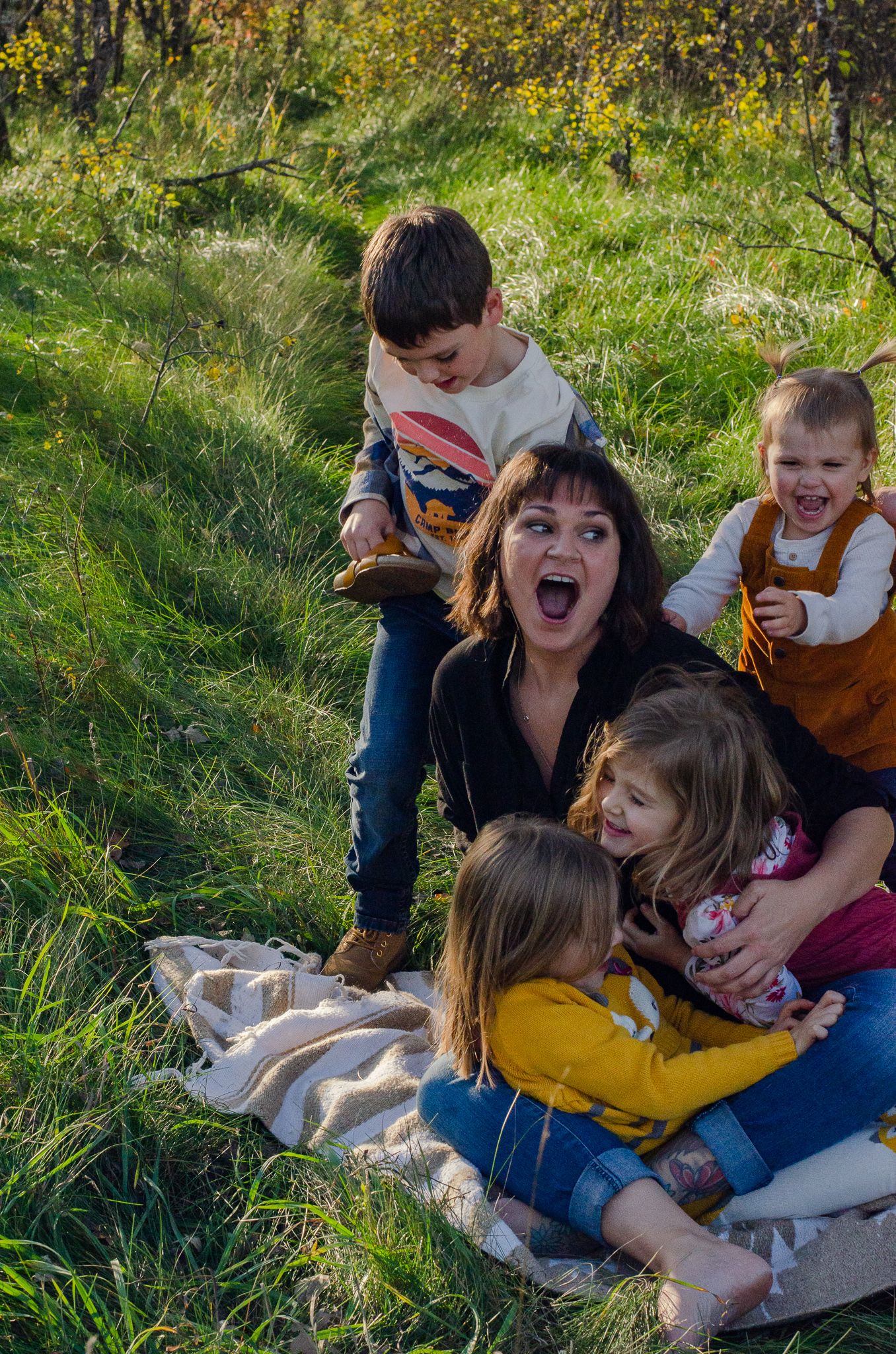 A joyful mother sits on a blanket in a green field laughing with her four young children climbing around her.