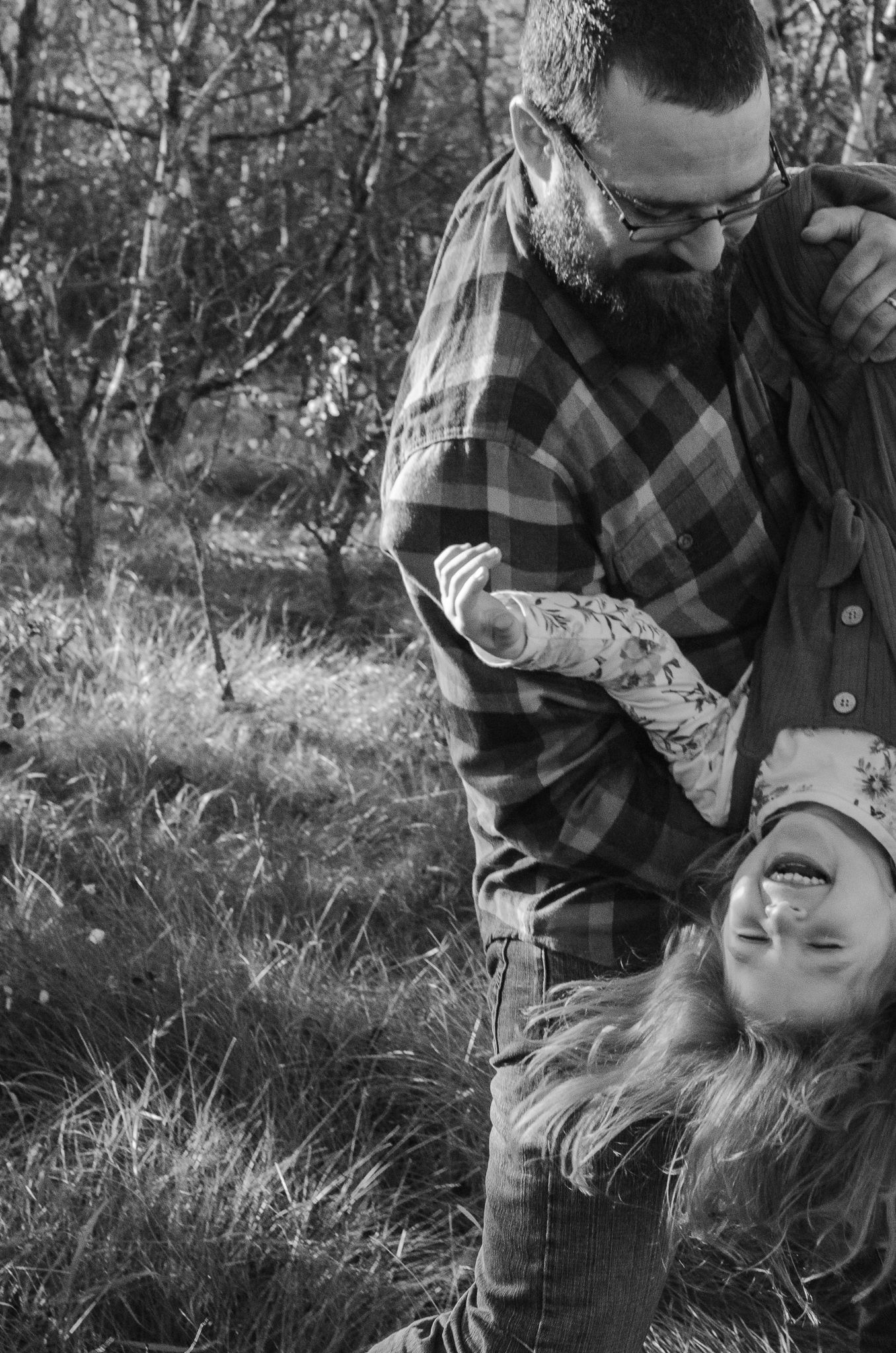 Black and white photo of a bearded father playfully holding his laughing child upside down outdoors.