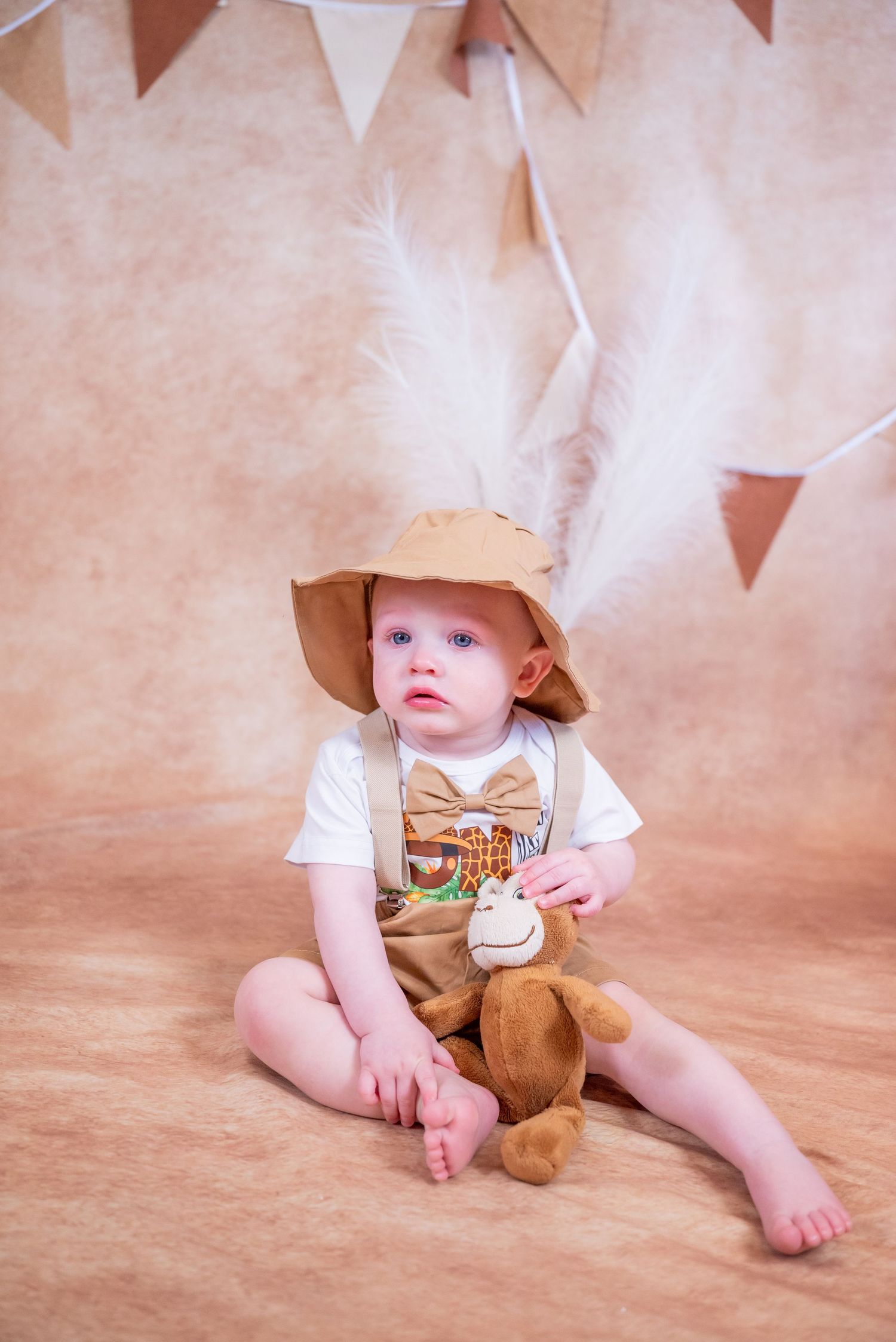 Baby boy in brown hat and suspenders sits on floor holding stuffed monkey toy with bunting decorations behind him.