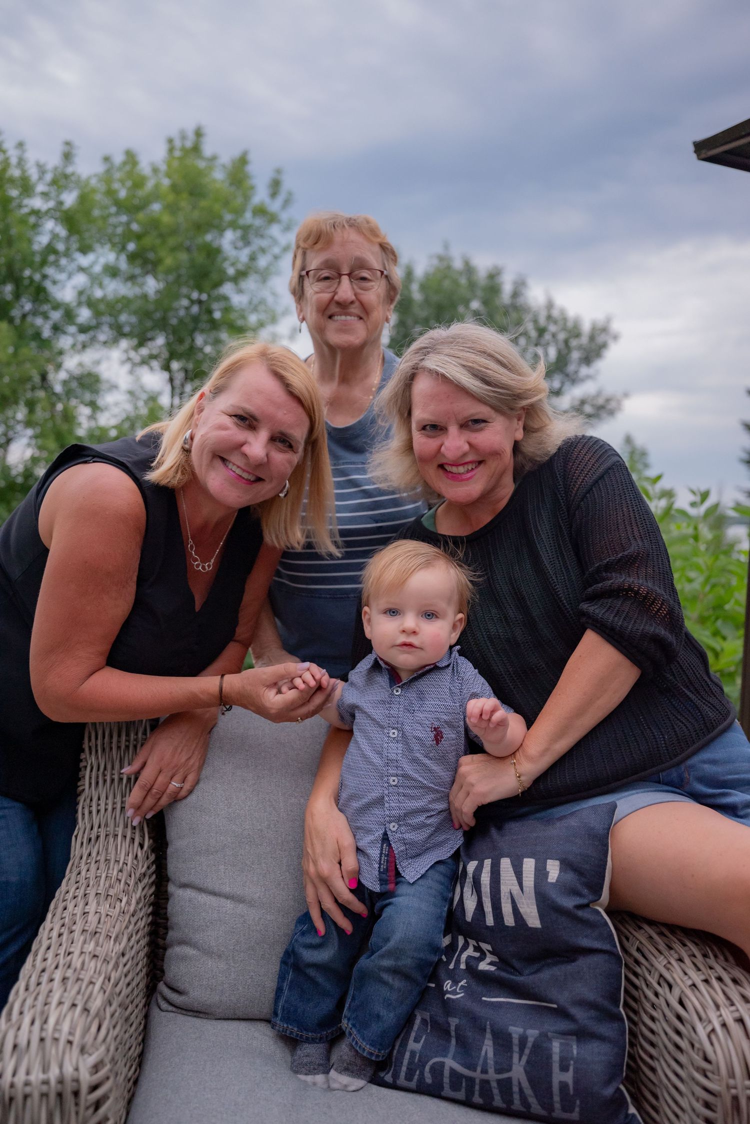 Three generations of women smiling outdoors with a baby boy in a gray shirt and jeans on a cloudy day.
