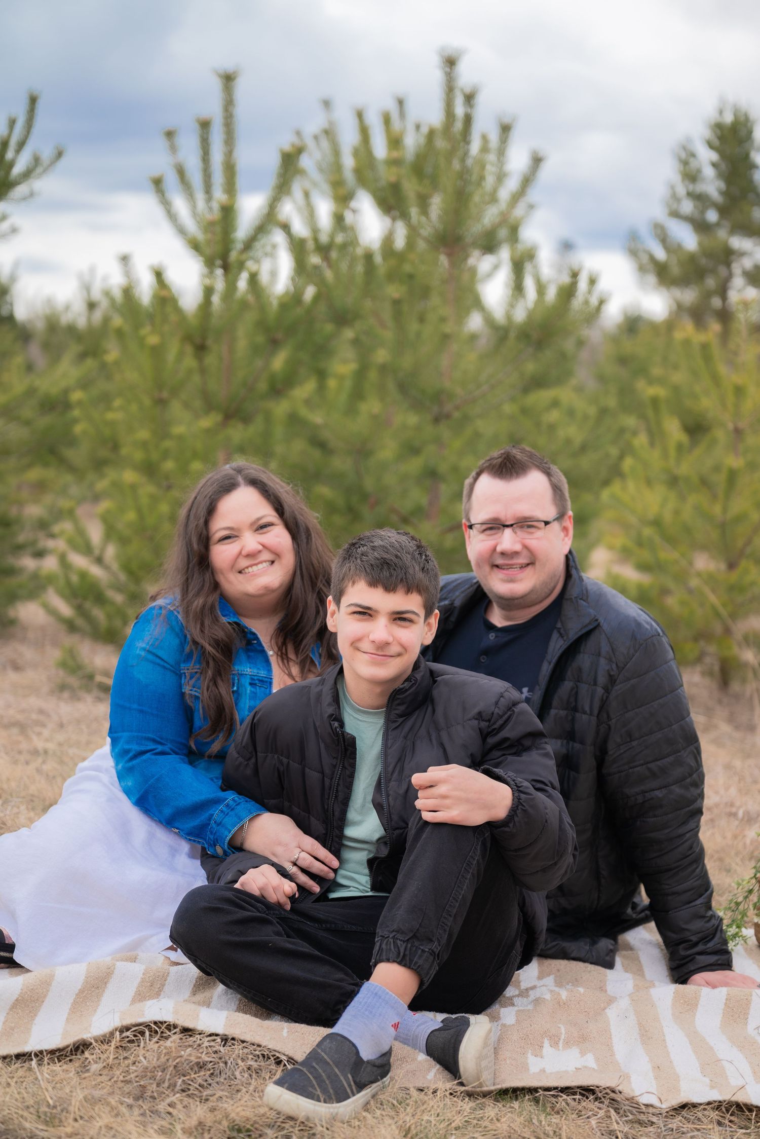 Family of three smiling together on a blanket outdoors, surrounded by pine trees on a cloudy day.