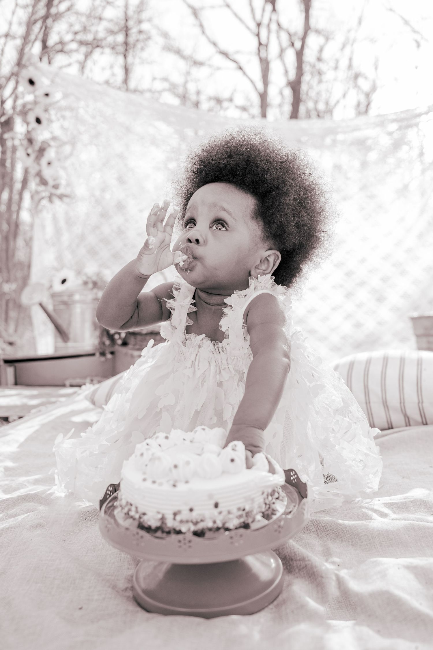 Black and white photo of a baby girl in white lace dress eating birthday cake at her first birthday party.