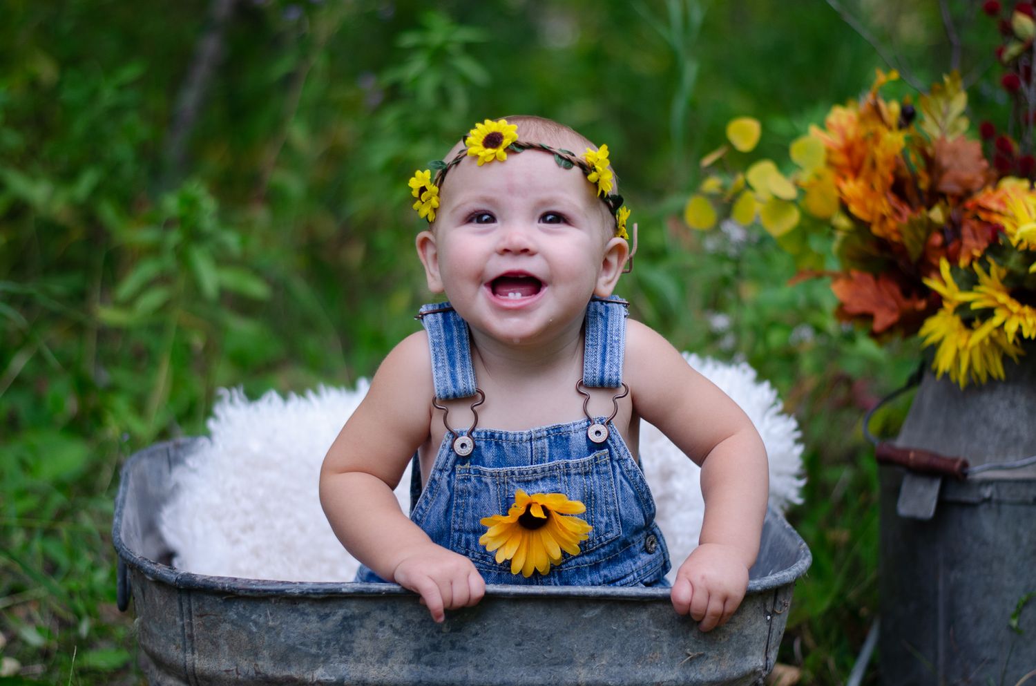 Smiling baby in denim overalls with sunflower accessories sitting in a vintage metal tub outdoors.