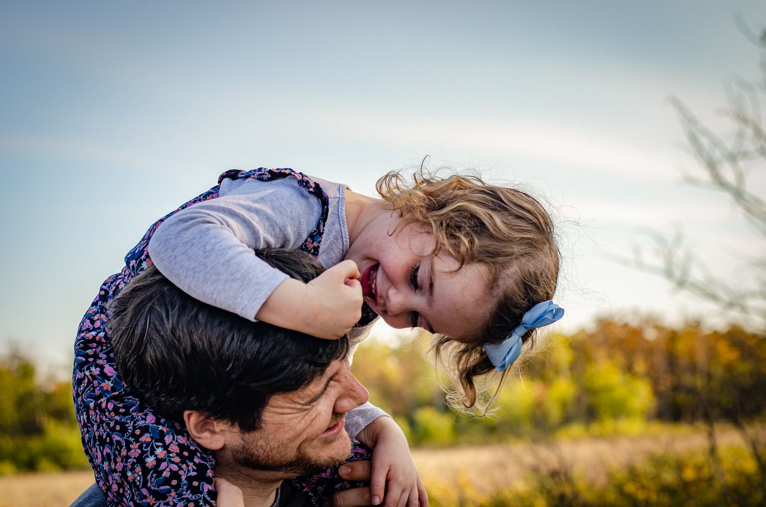 A joyful young girl kisses a woman on the head while being carried on her shoulders outdoors.