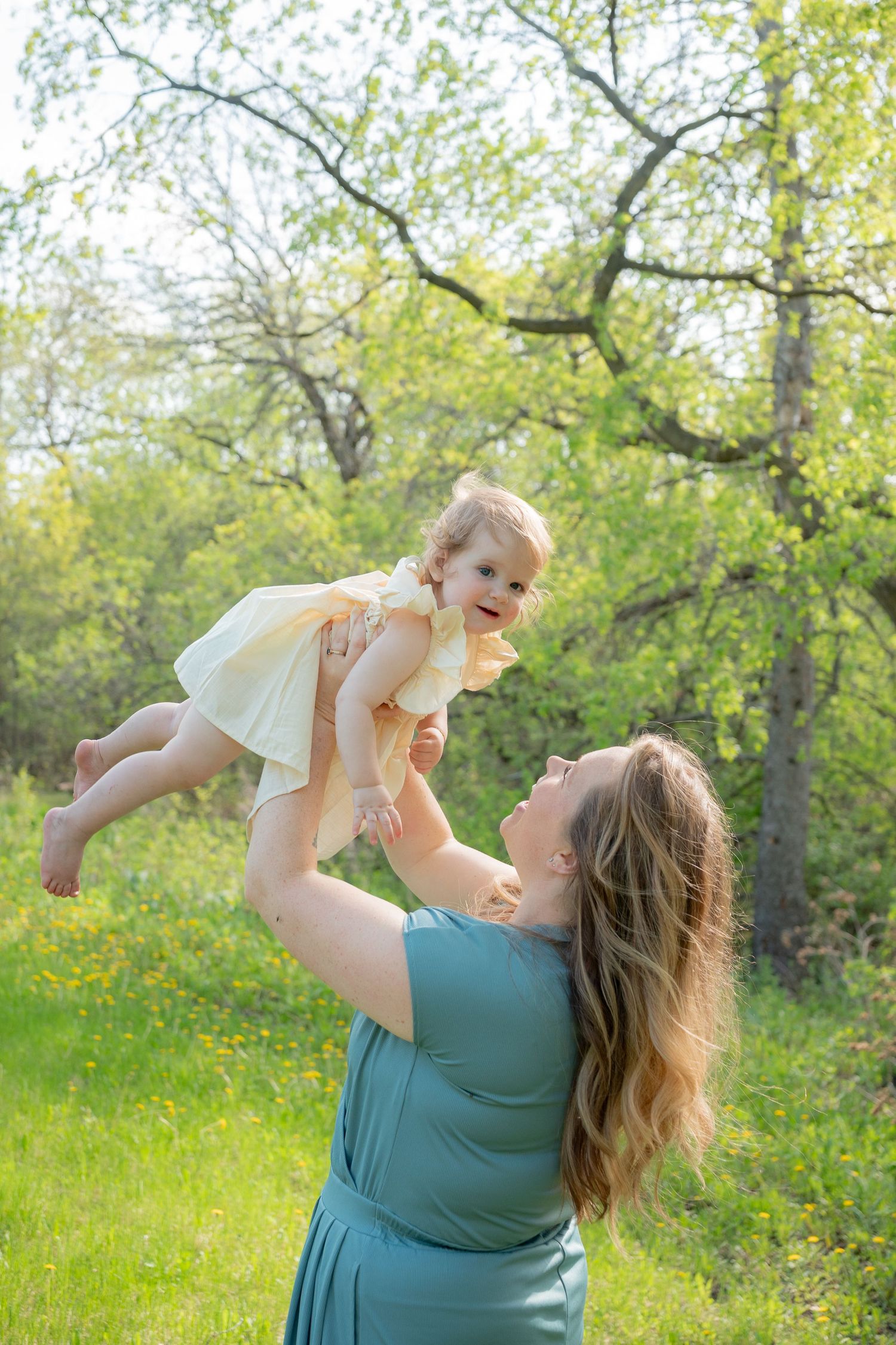 A joyful mother in a teal dress lifts her smiling baby girl in a yellow dress outdoors in a lush green park.