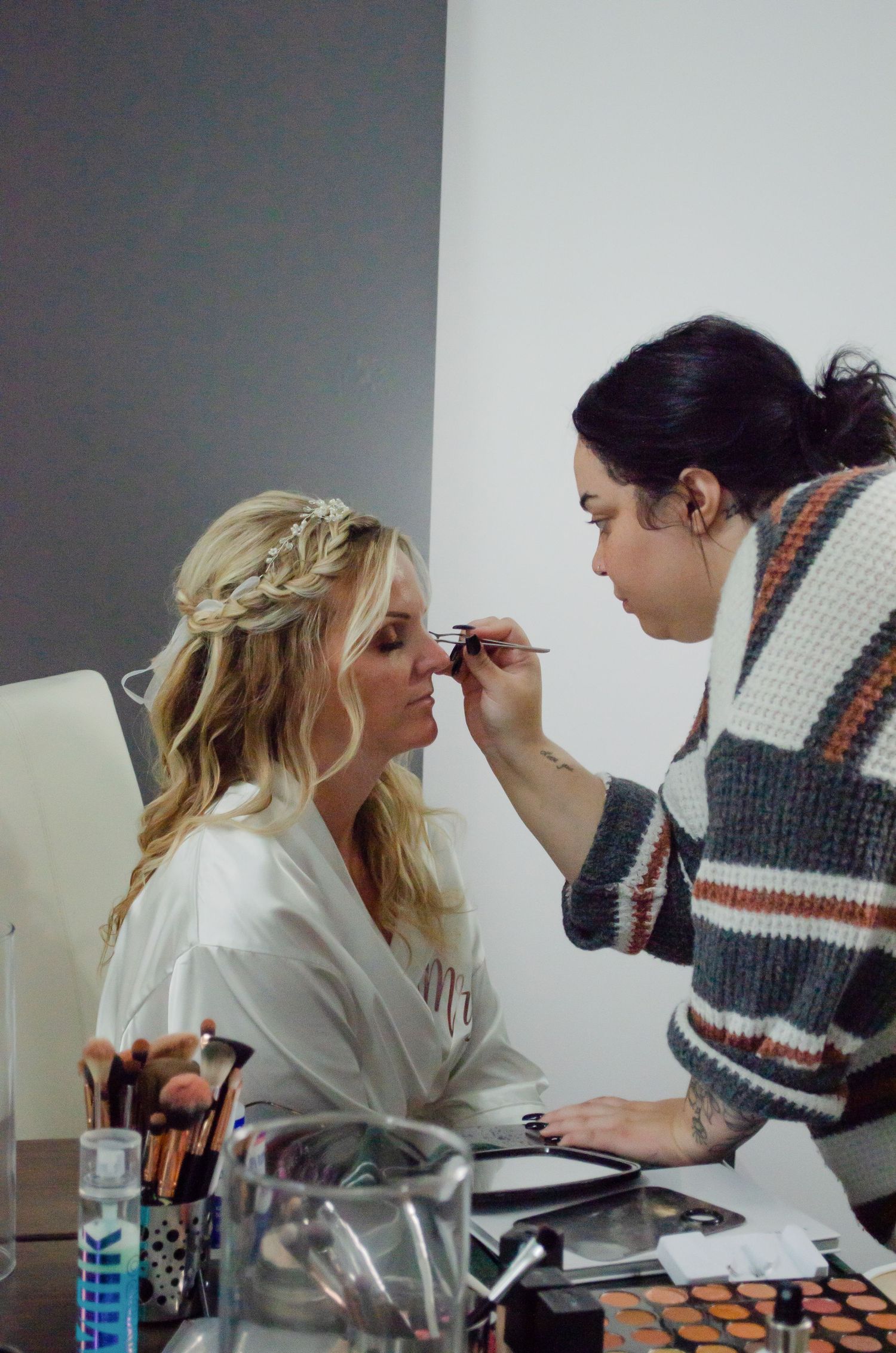 A makeup artist applying eye makeup to a blonde bride with a braided updo, wearing a white robe, at a vanity.