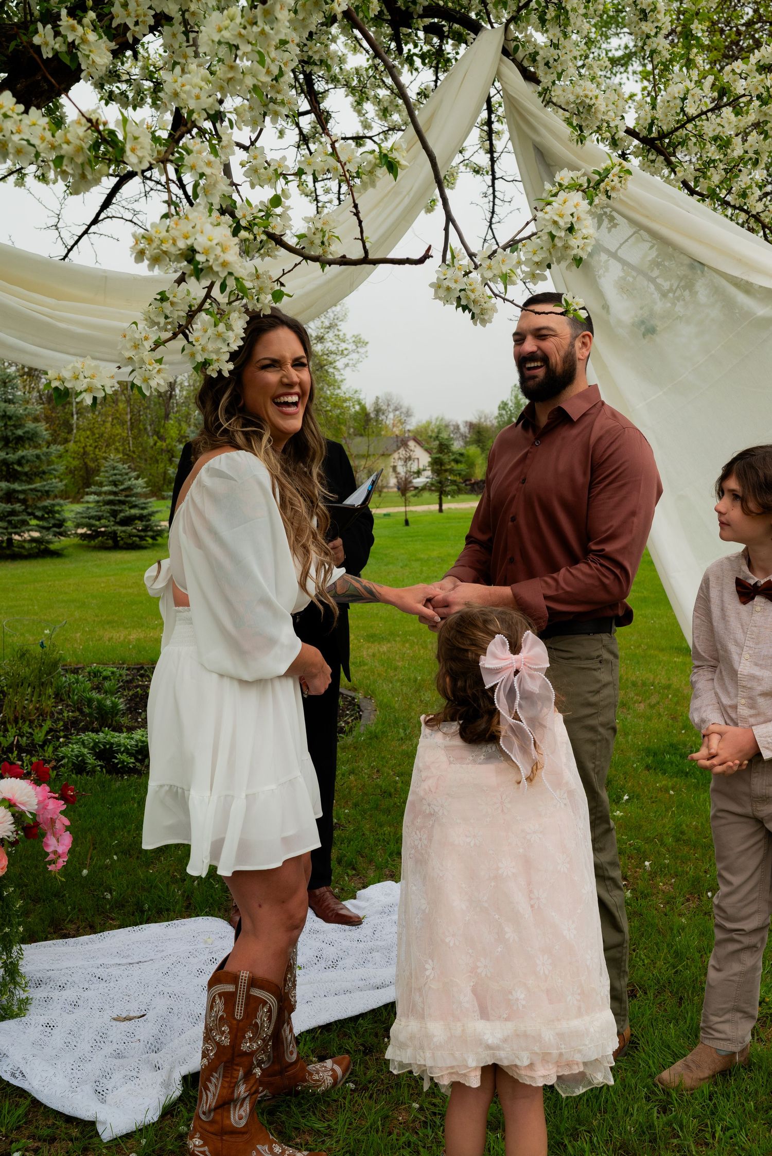 Couple exchanging vows at outdoor wedding ceremony under blooming tree with flower girl and boy nearby.