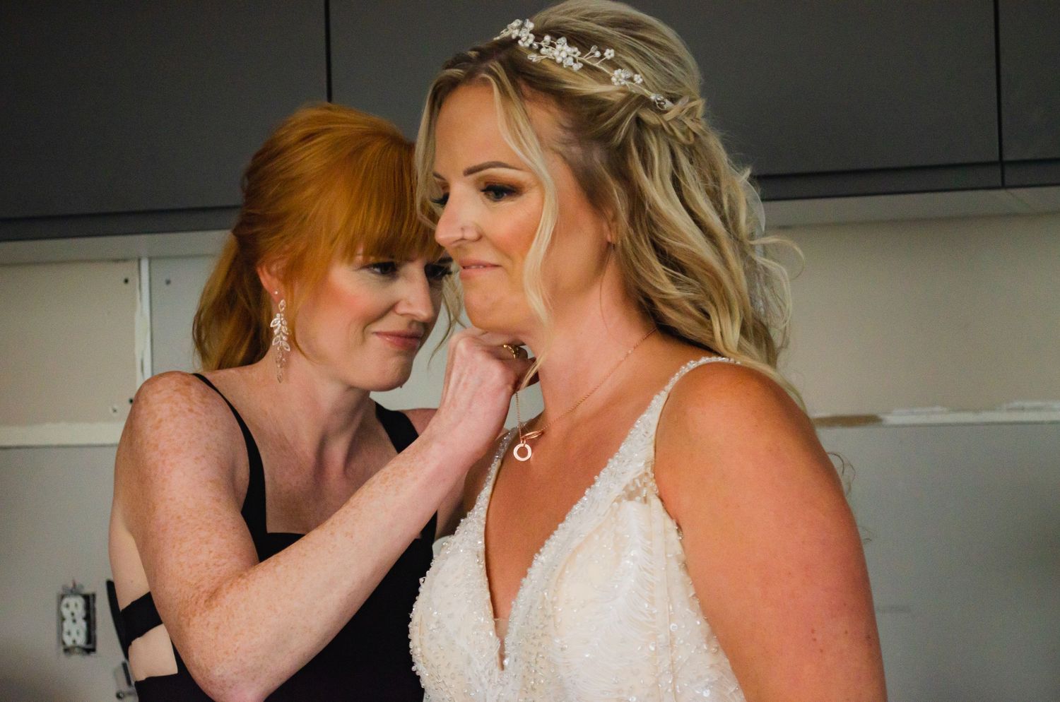 A redhead bridesmaid adjusts the necklace of a blonde bride in a white lace gown wearing a floral hair accessory.