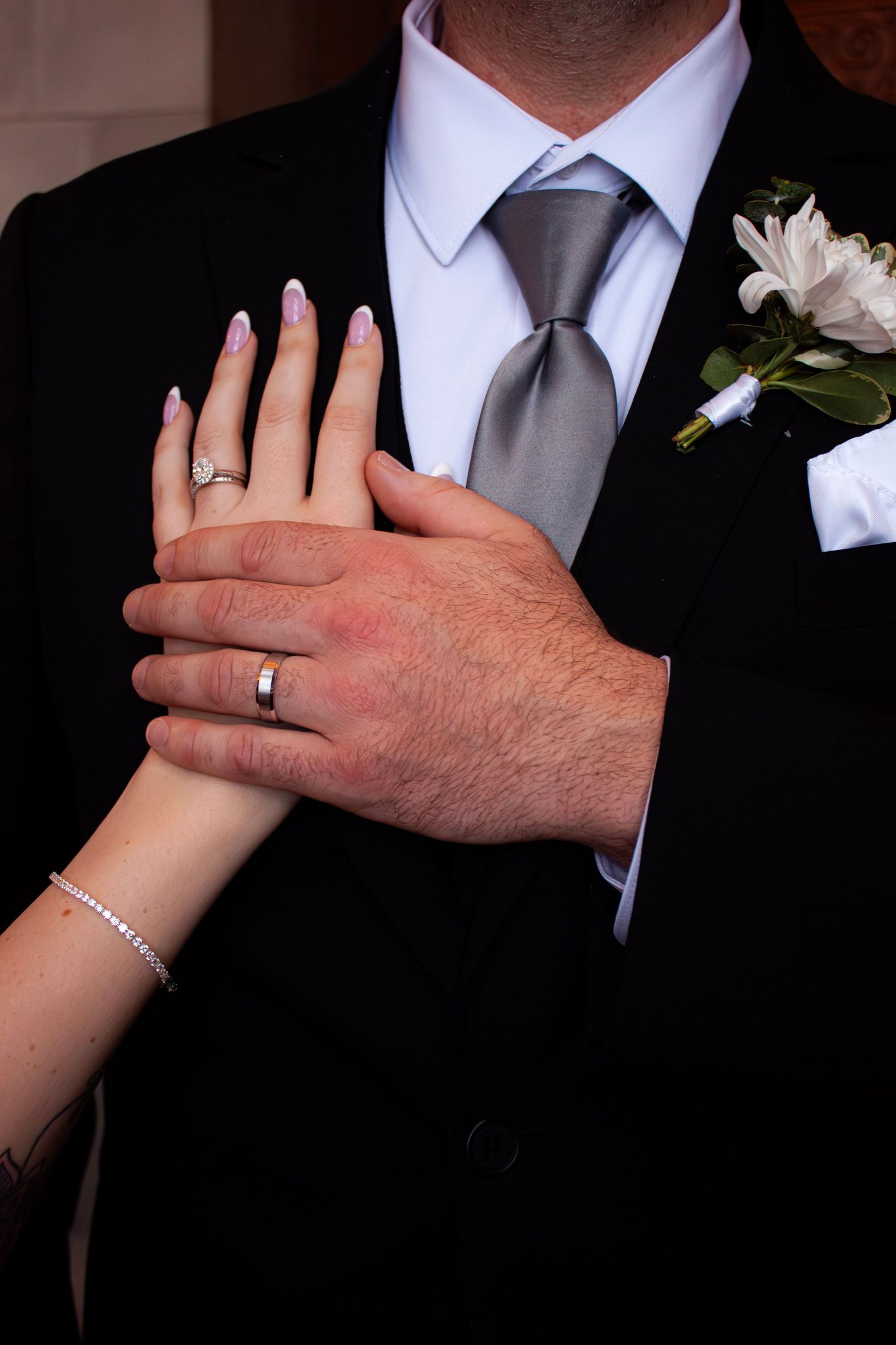 Bride and groom displaying wedding rings, her hand resting on his chest in black suit with gray tie and white boutonniere.
