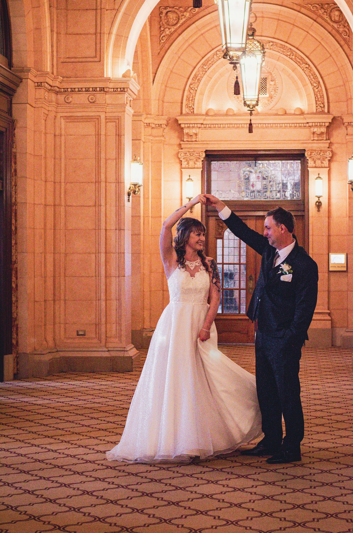 Bride and groom share a romantic dance in an elegant golden-lit ballroom hallway on their wedding day.