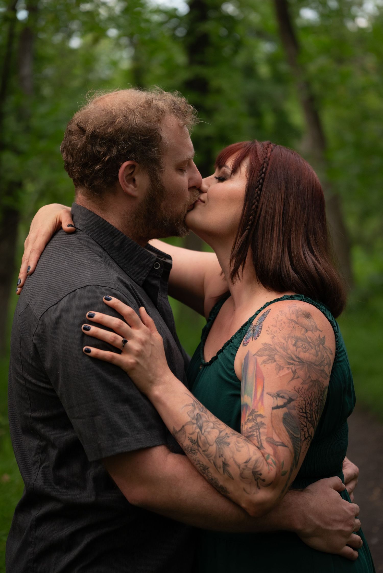 Couple kissing in a lush green forest, woman with tattoo sleeve in green dress embracing man in black shirt.