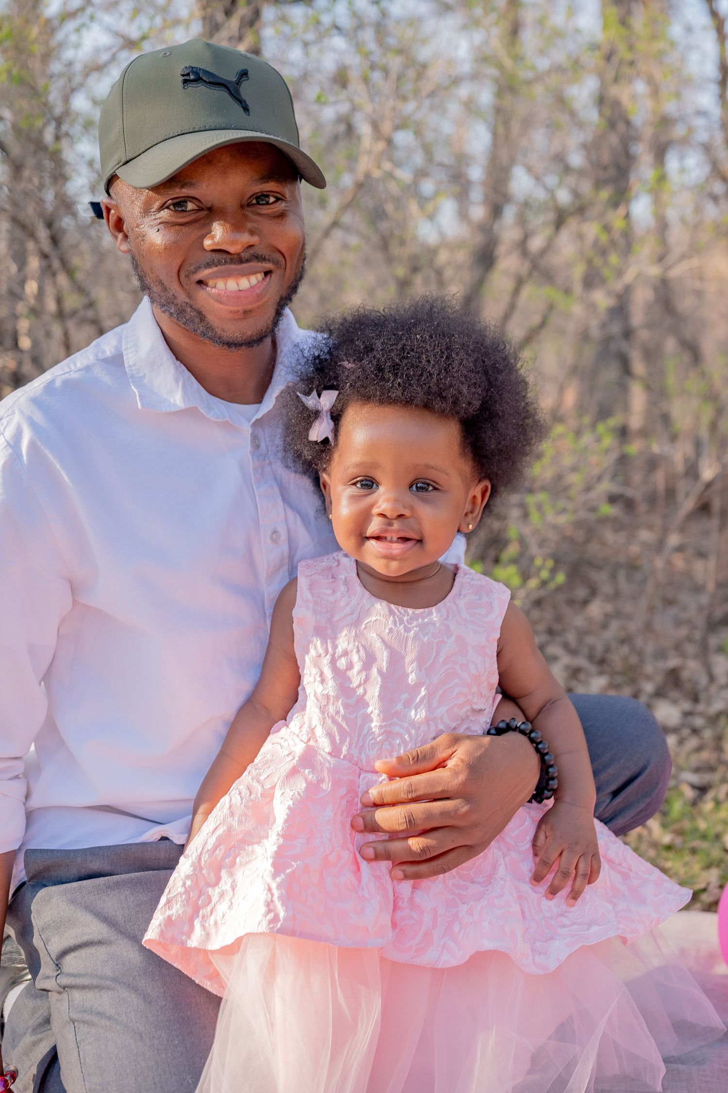 Smiling father in Puma cap holds young daughter in pink dress outdoors.
