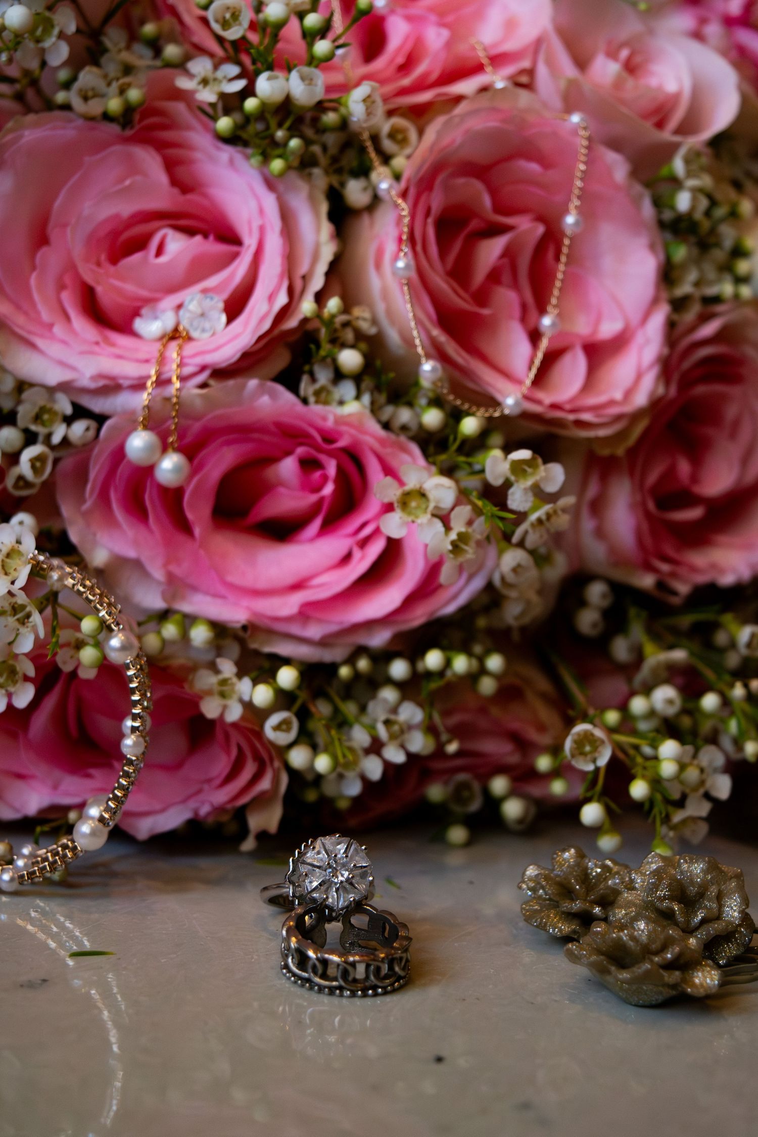Close-up of wedding rings beside pink roses bouquet with pearl bracelet on marble surface.