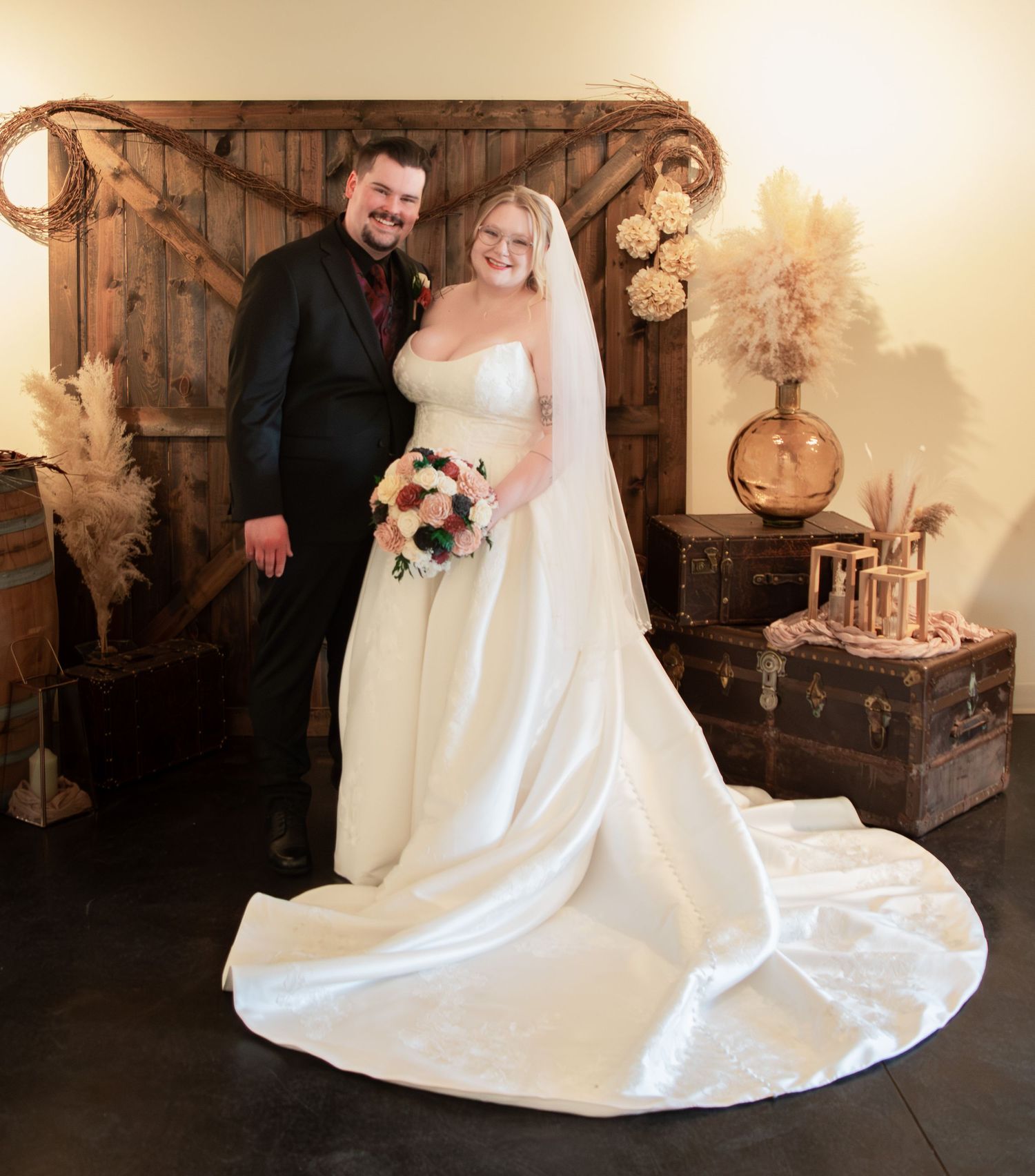 Bride and groom pose together before rustic wooden barn door backdrop with pampas grass decor.