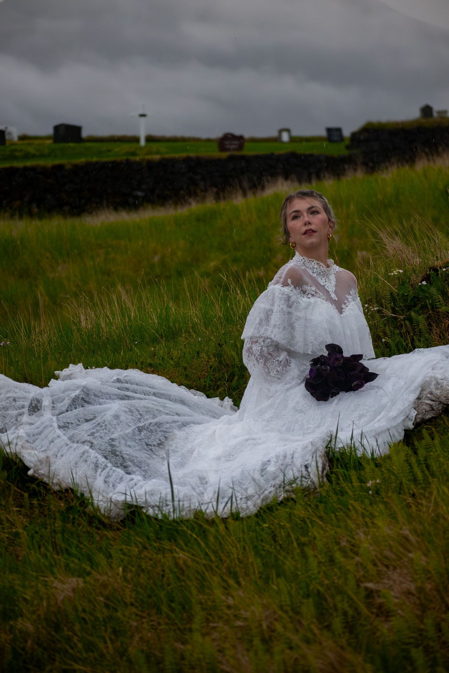 Bride in elegant lace wedding gown sits gracefully in green meadow holding dark purple bouquet under cloudy sky.
