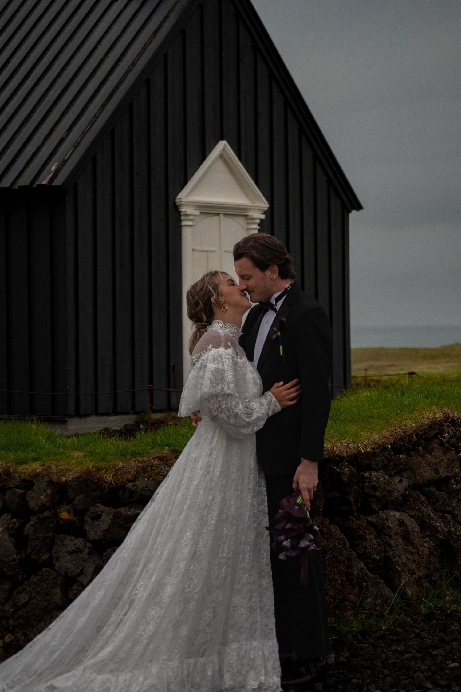 Bride and groom share a romantic moment outside a black wooden church in Iceland, bride wearing a flowing lace gown.