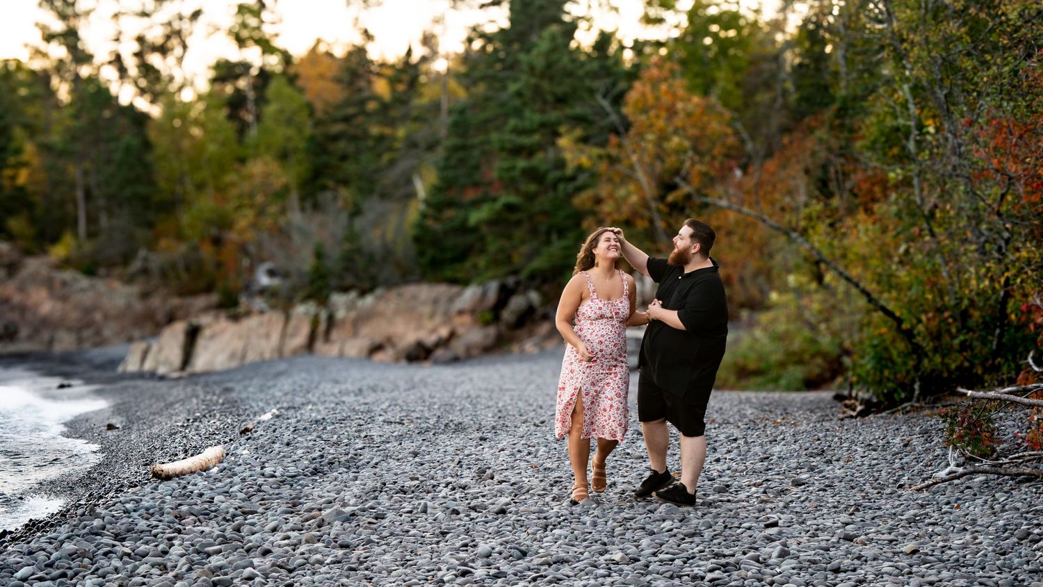 Happy couple walking hand-in-hand along a rocky beach surrounded by autumn foliage.